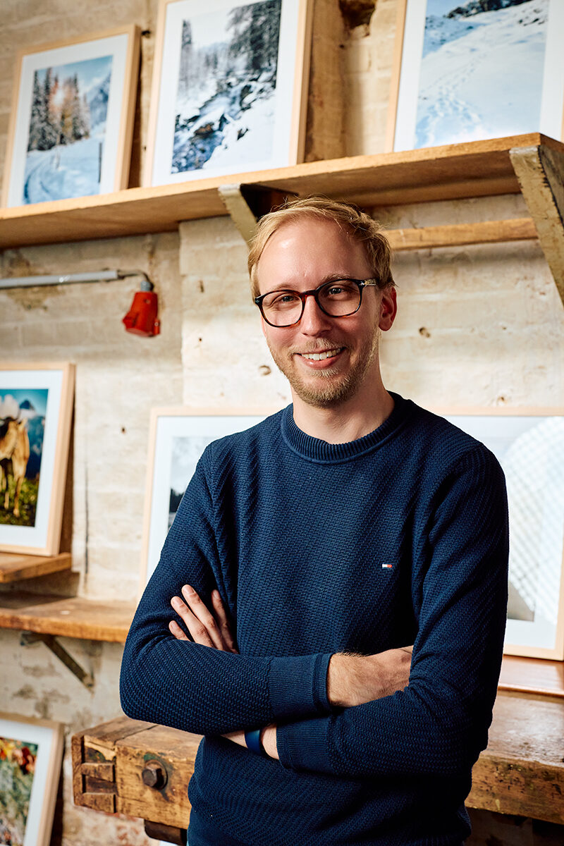 Portrait von Fotograf Christopher Fuhrmann mit Bildern seiner Ausstellung Walser Stories im Hintergrund.