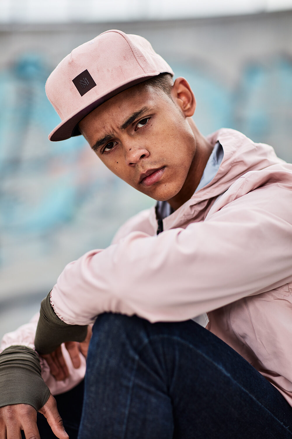 Color-Block-Streetwear-Editorial: Nahaufnahme von Model David mit rosa Cap und Anorak vor einer Graffiti-Wand.