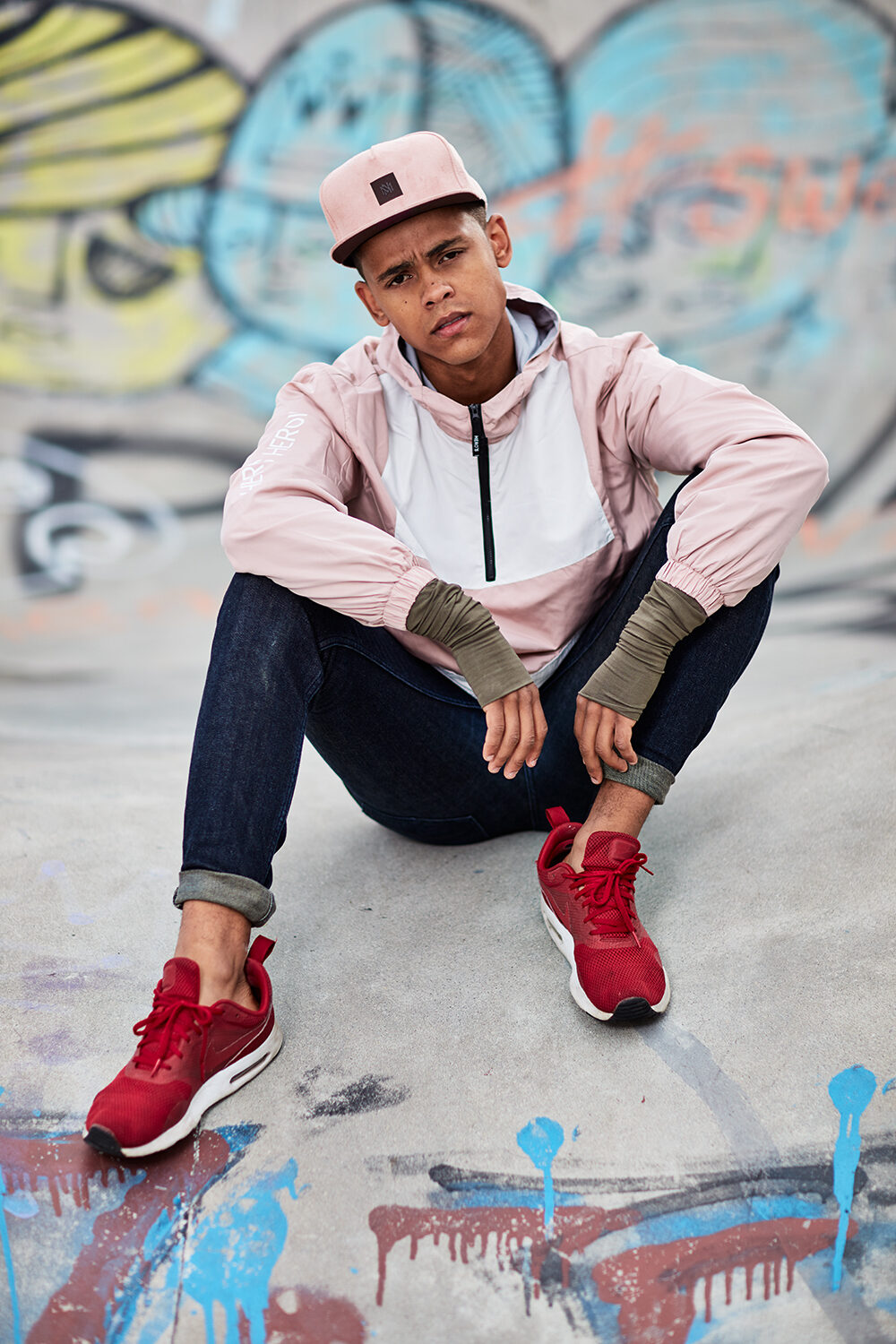 Streetwear-Editorial: Model David mit rosa Cap und Anorak sitzt vor einer Graffiti-Wand im Skatepark.