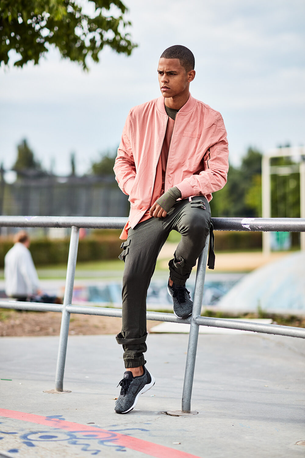 Streetwear-Editorial: Model David in lachsrosa Bomberjacke und Cargohose sitzt auf einem Geländer im Skatepark.