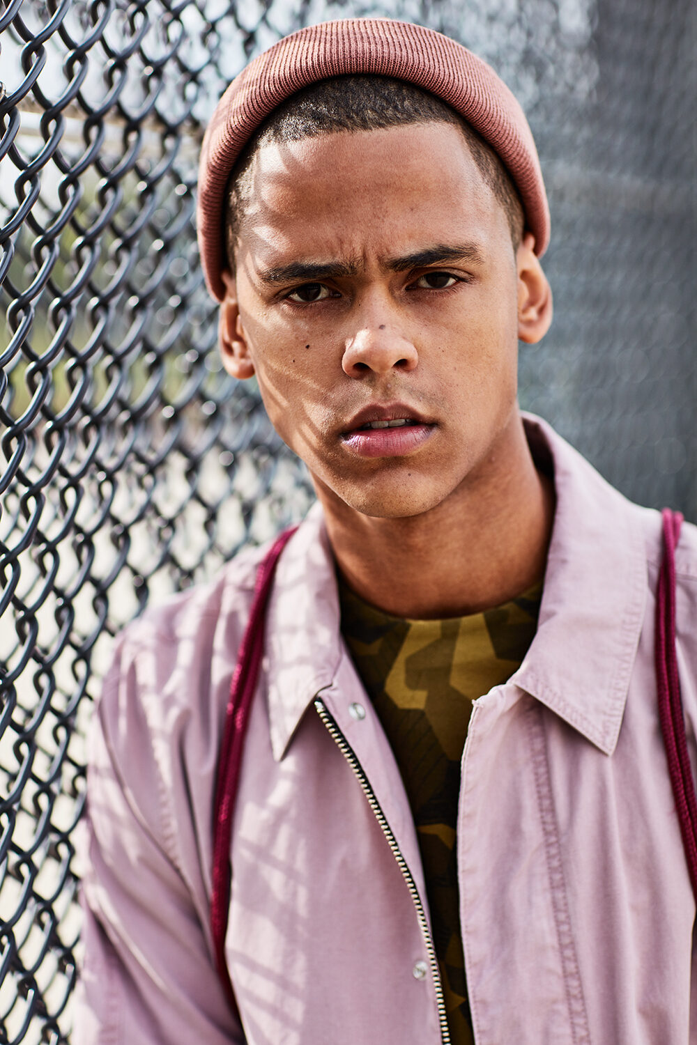 Streetwear-Editorial: Model David mit rosa Mütze und Hemd über Camouflage-Shirt vor einem Maschendrahtzaun.