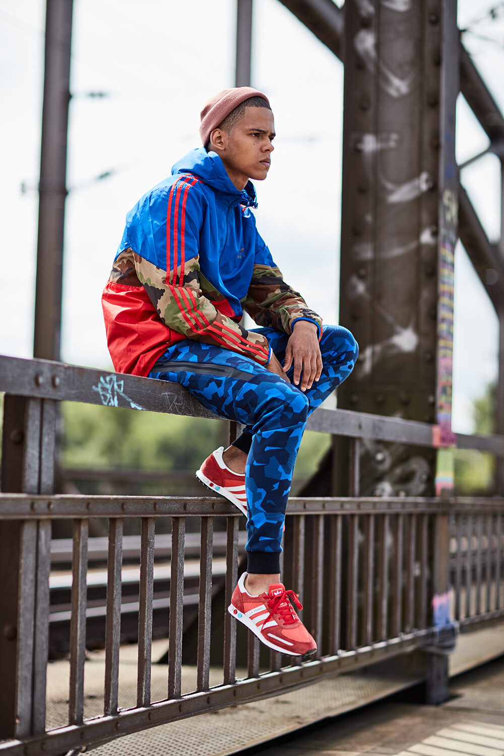 Color-Block-Editorial: Model David in blauem Adidas-Hoodie und Camouflage sitzt auf einem Brückengeländer.