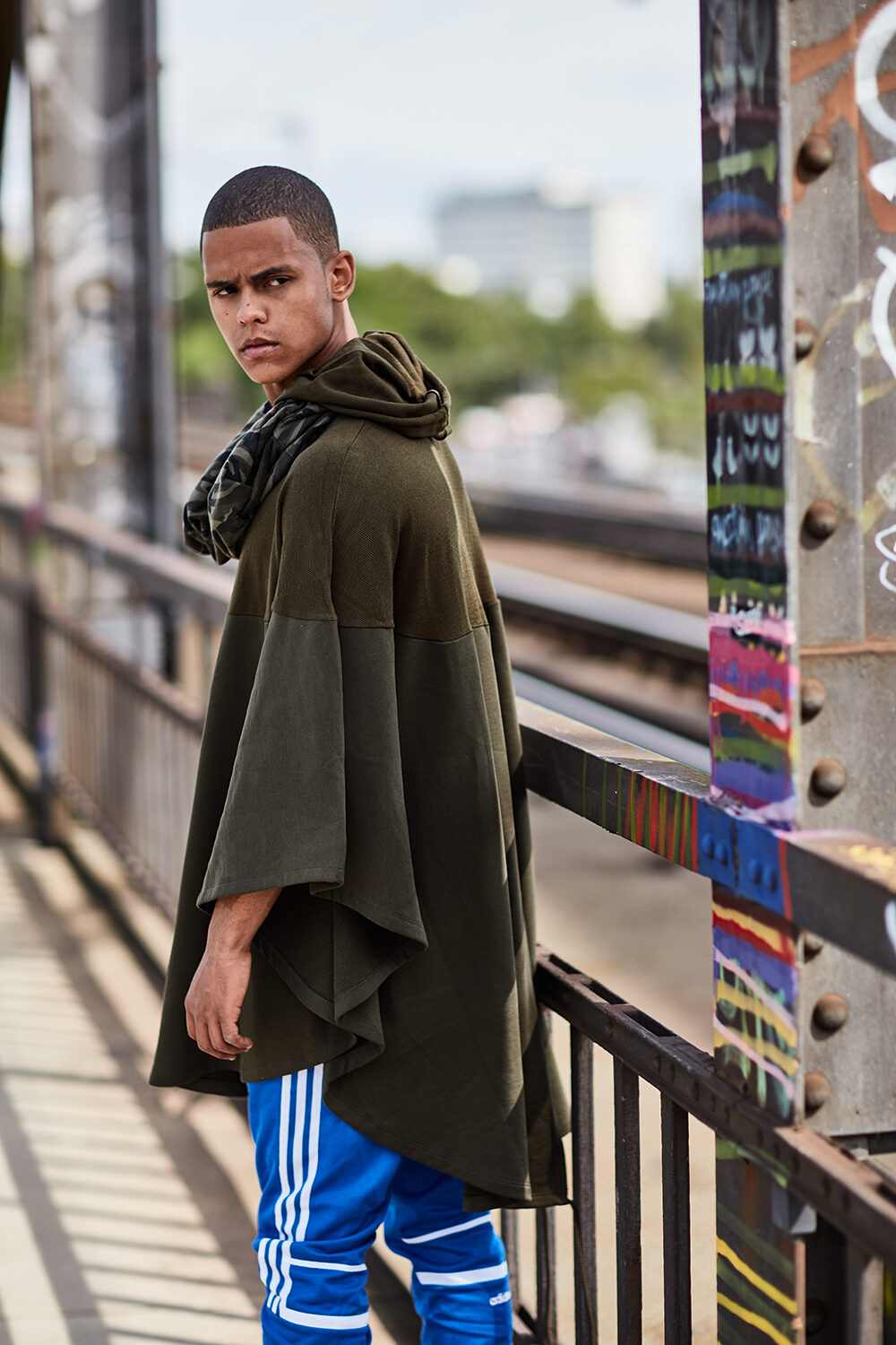 Streetwear-Editorial: Model David im olivgrünen Cape-Hoodie und blauer Trainingshose auf einer Graffiti-Brücke.