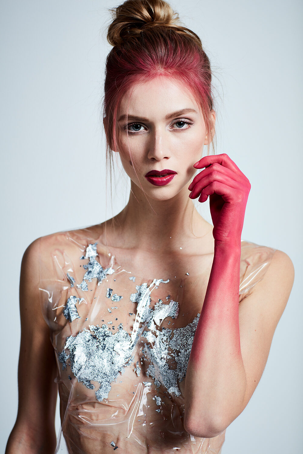 Konzeptionelle Beauty-Aufnahme: Frau mit pink gefärbtem Haar, in Klarsichtfolie und Silberblatt gehüllt, eine Hand pink bemalt.