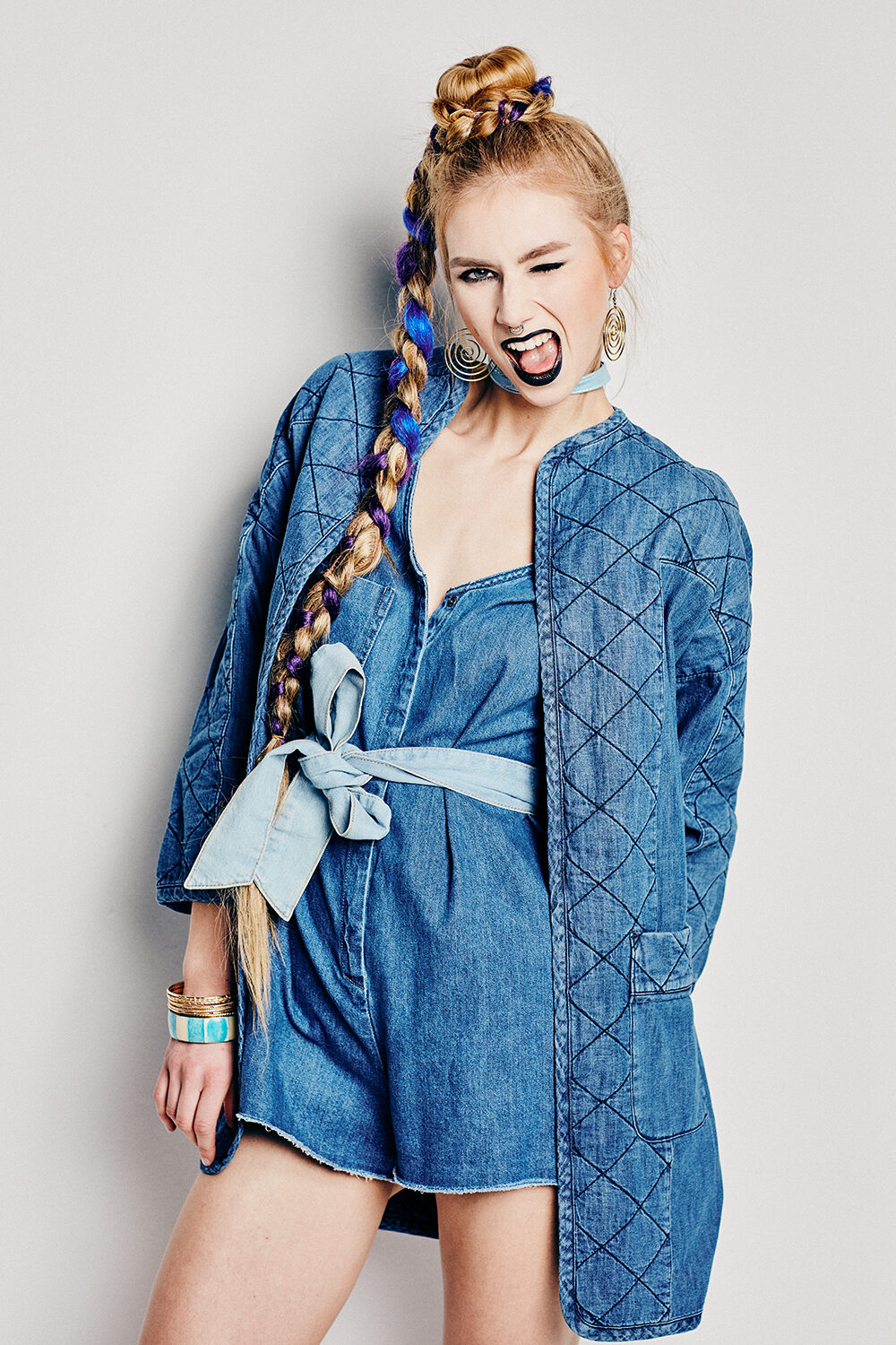 Denim-Editorial: blonde Frau mit blau geflochtenem Zopf in Steppjeansjacke über Jeans-Playsuit vor hellem Hintergrund.