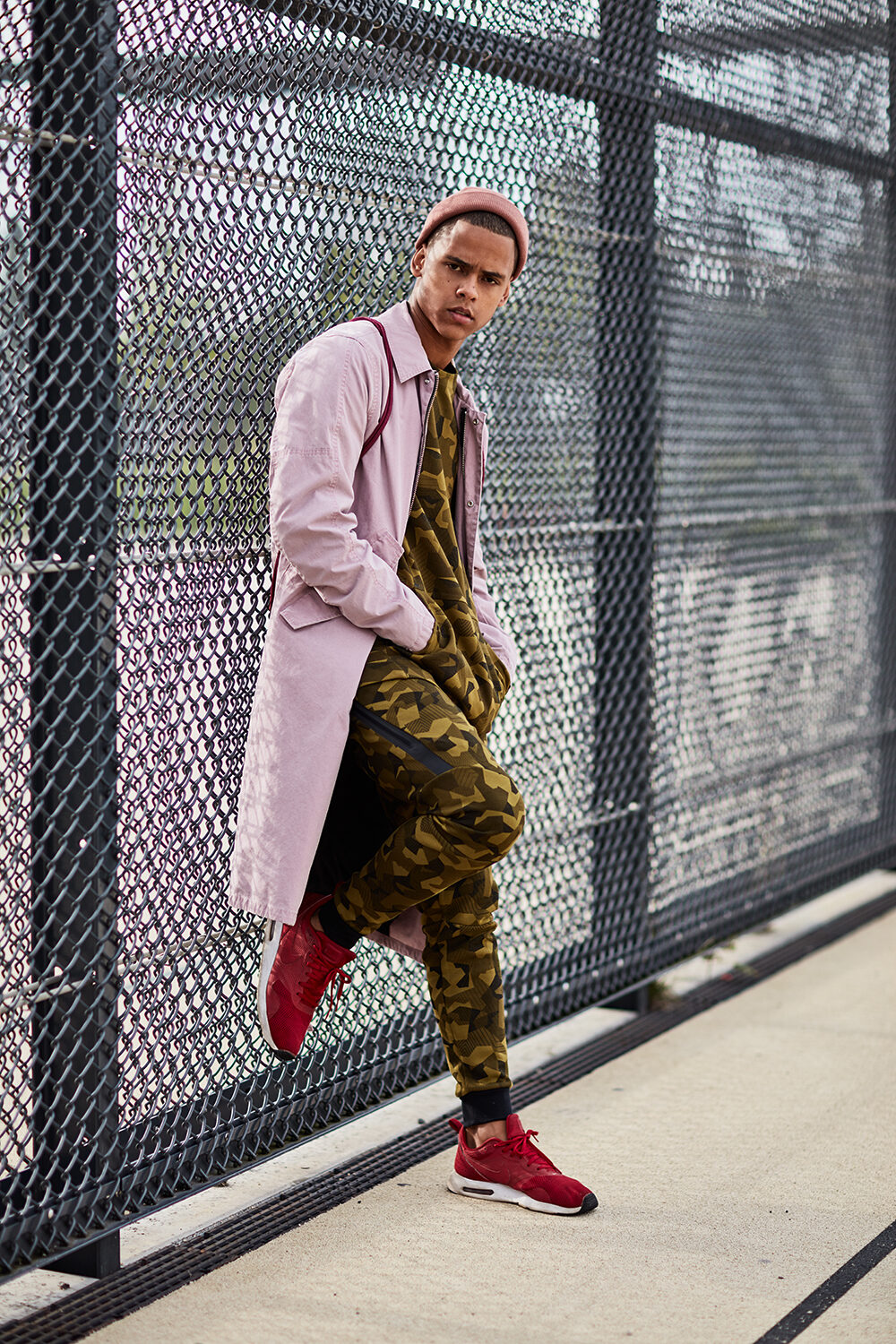 Streetwear-Editorial: männliches Model im rosa Mantel über Camouflage-Overall vor einem Maschendrahtzaun.