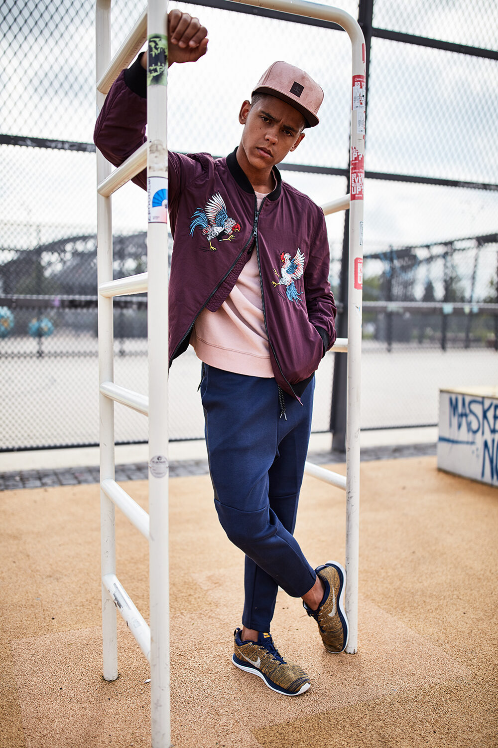 Streetwear-Editorial: männliches Model in Bomberjacke mit Kranich-Stickerei an einem Basketballkäfig.