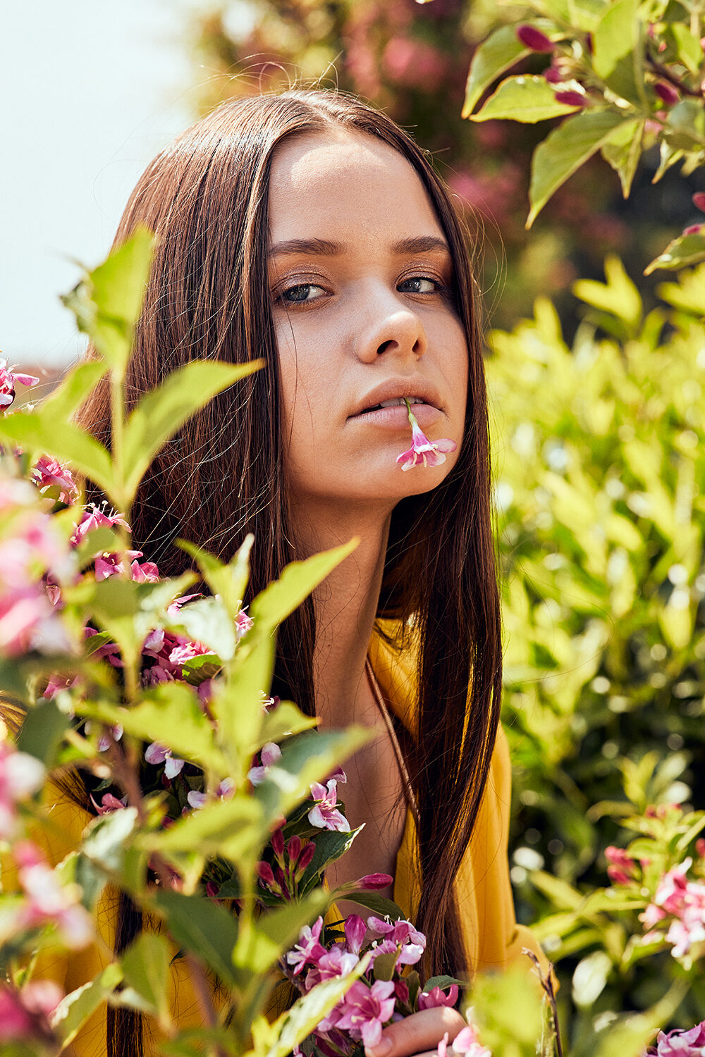 Beauty-Editorial im Freien: brünette Frau in Gelb zwischen rosa Blüten, eine Blüte an den Lippen.