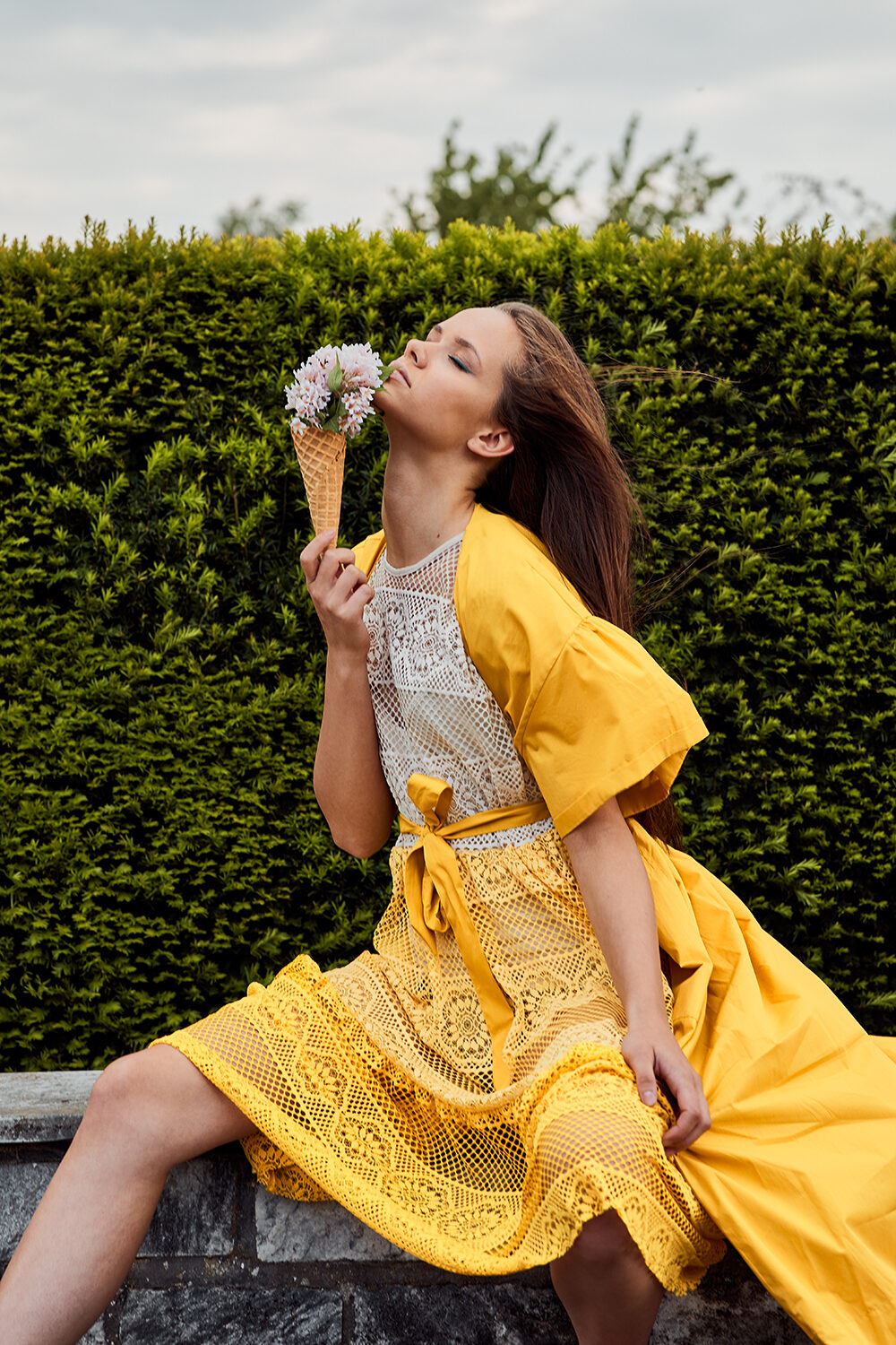Konzept-Editorial: Frau im gelben Kleid hält eine mit Blüten gefüllte Eiswaffel, sitzend vor einer Hecke.