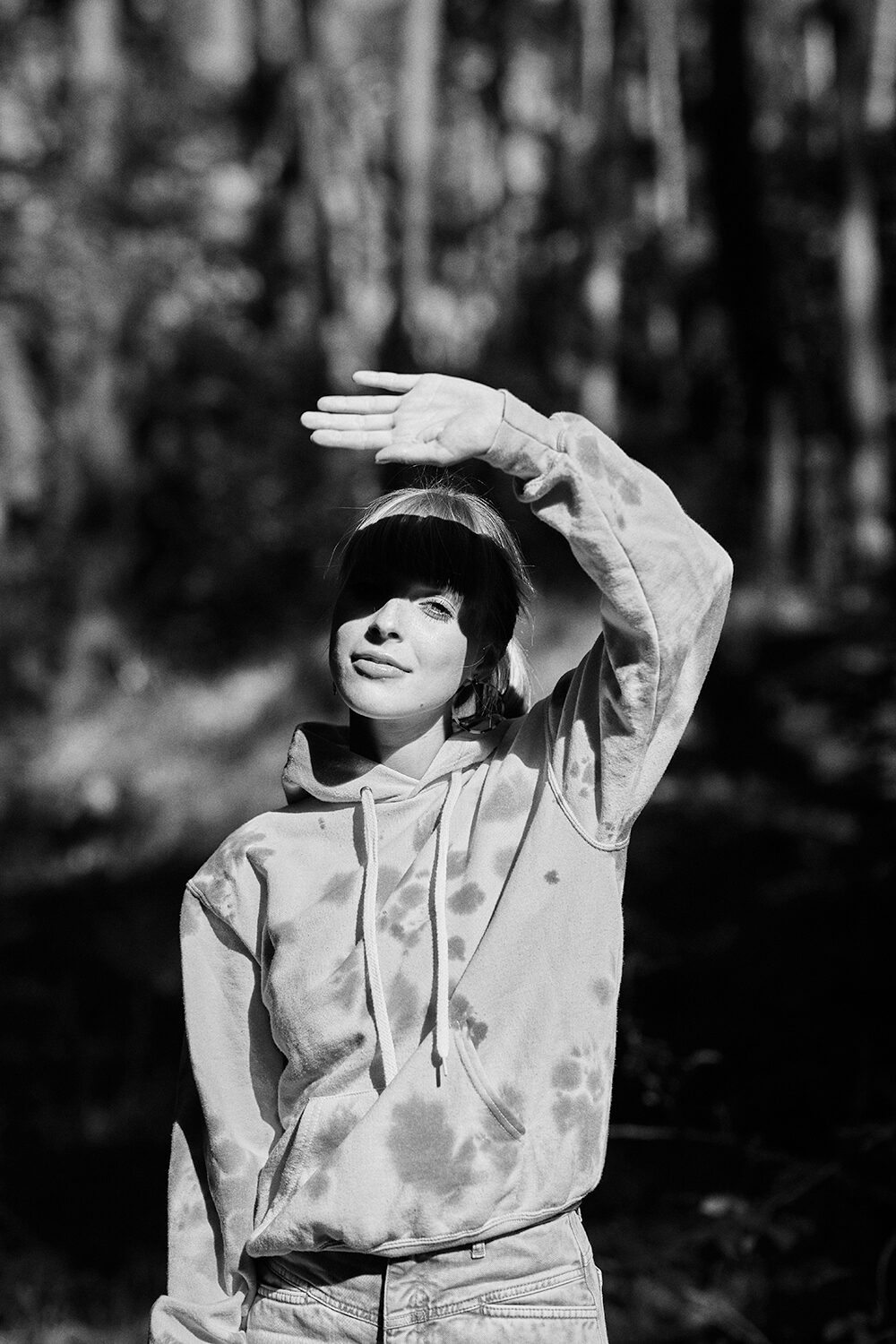 Schwarz-Weiß-Editorial: Frau im Batik-Hoodie schirmt im Wald die Augen mit der Hand ab.