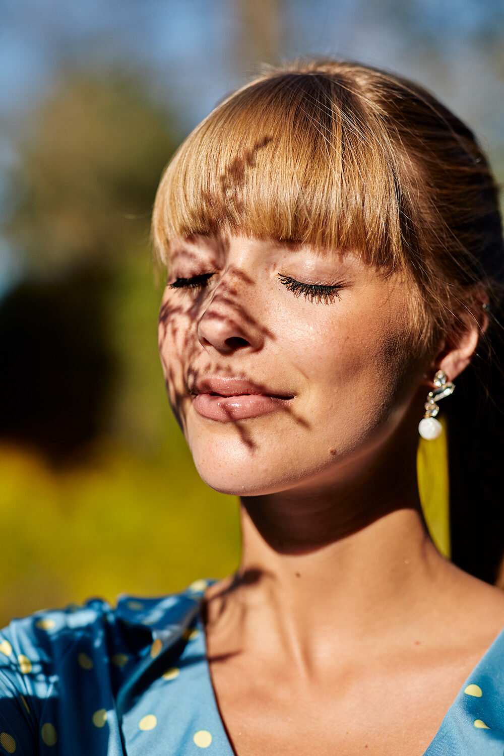 Beauty-Editorial: Nahaufnahme einer blonden Frau mit Pony, geschlossenen Augen und Blätterschatten im Gesicht.