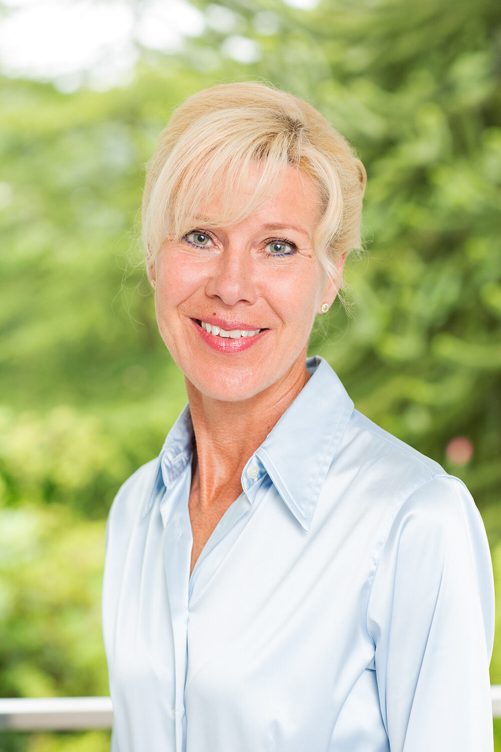 Businessportrait einer lächelnden Frau in hellblauer Bluse vor grünem, unscharfem Hintergrund.