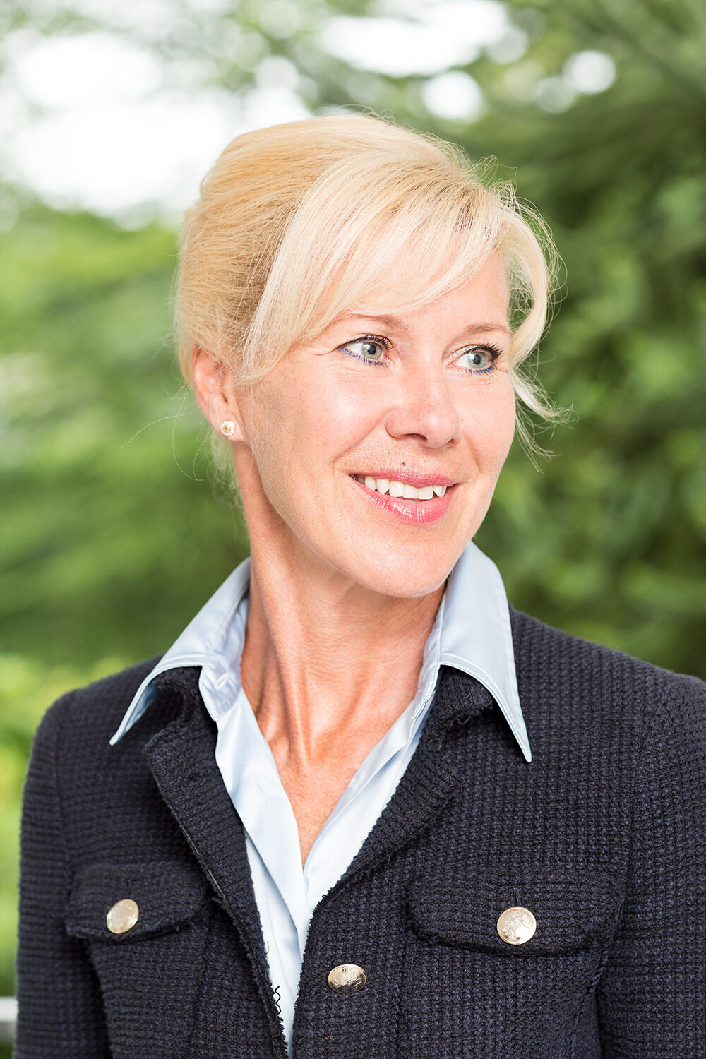 Businessportrait einer lächelnden Frau im dunklen Tweed-Blazer über hellblauer Bluse vor grünem, unscharfem Hintergrund.