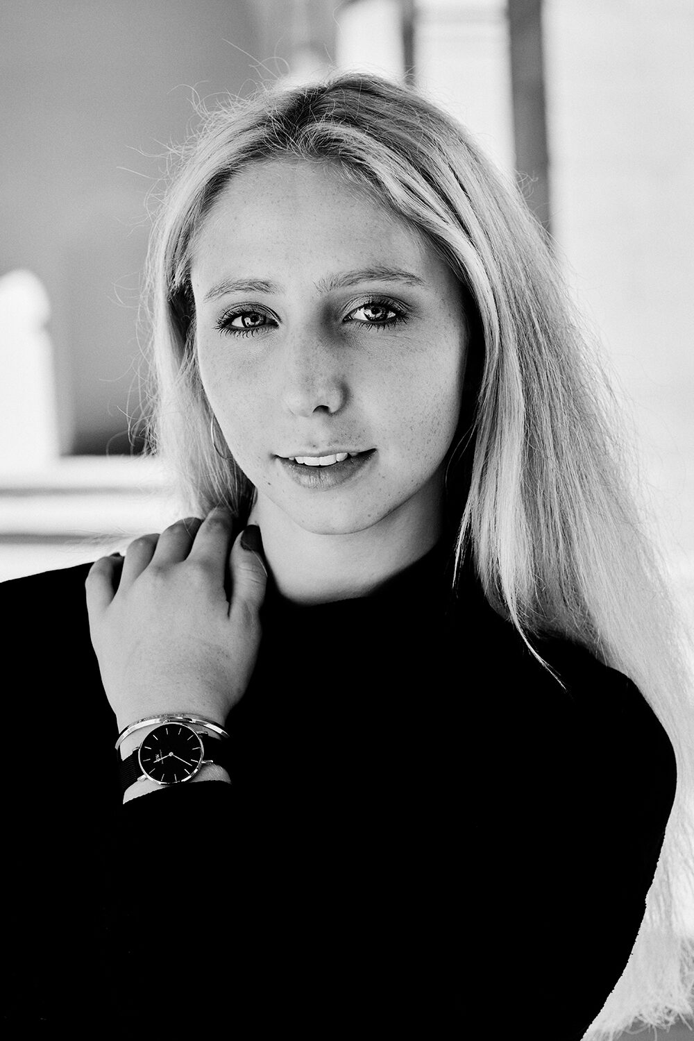 Schwarz-Weiß-Portrait einer jungen blonden Frau im schwarzen Rollkragenpullover, die Hand mit Armbanduhr am Hals.