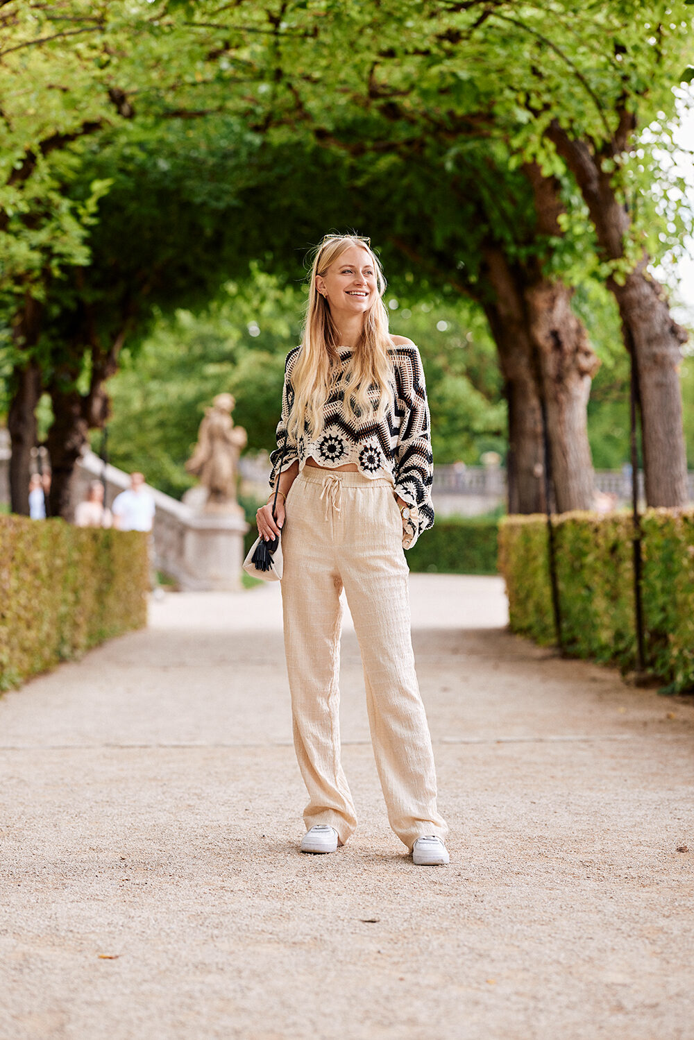 Blonde Frau im gemusterten Häkelpullover und heller Leinenhose geht lachend durch eine Baumallee im Schlossgarten.