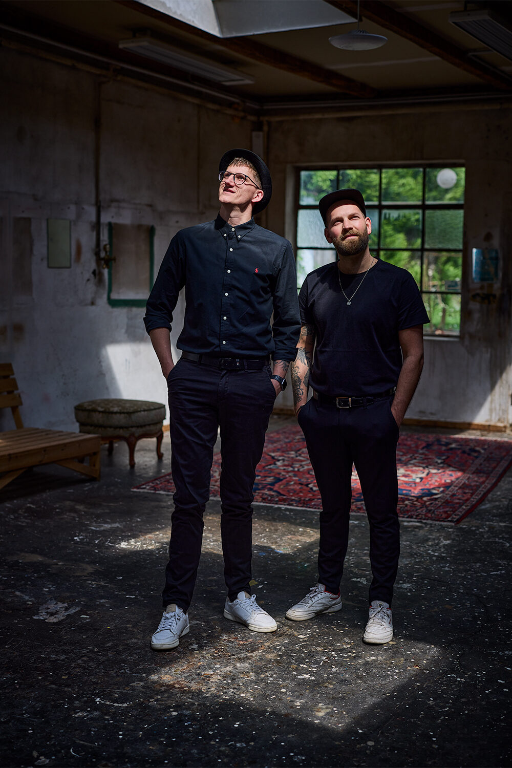 Zwei Männer in Schwarz stehen mit den Händen in den Taschen in einem leeren Industrieraum mit Teppich und altem Sofa und blicken zum Fenster.