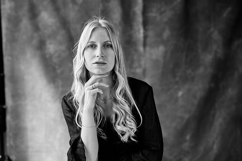 Schwarz-Weiß-Studioportrait einer Frau mit langem gewelltem Haar, die das Kinn in die Hand stützt, vor changierendem Hintergrund.