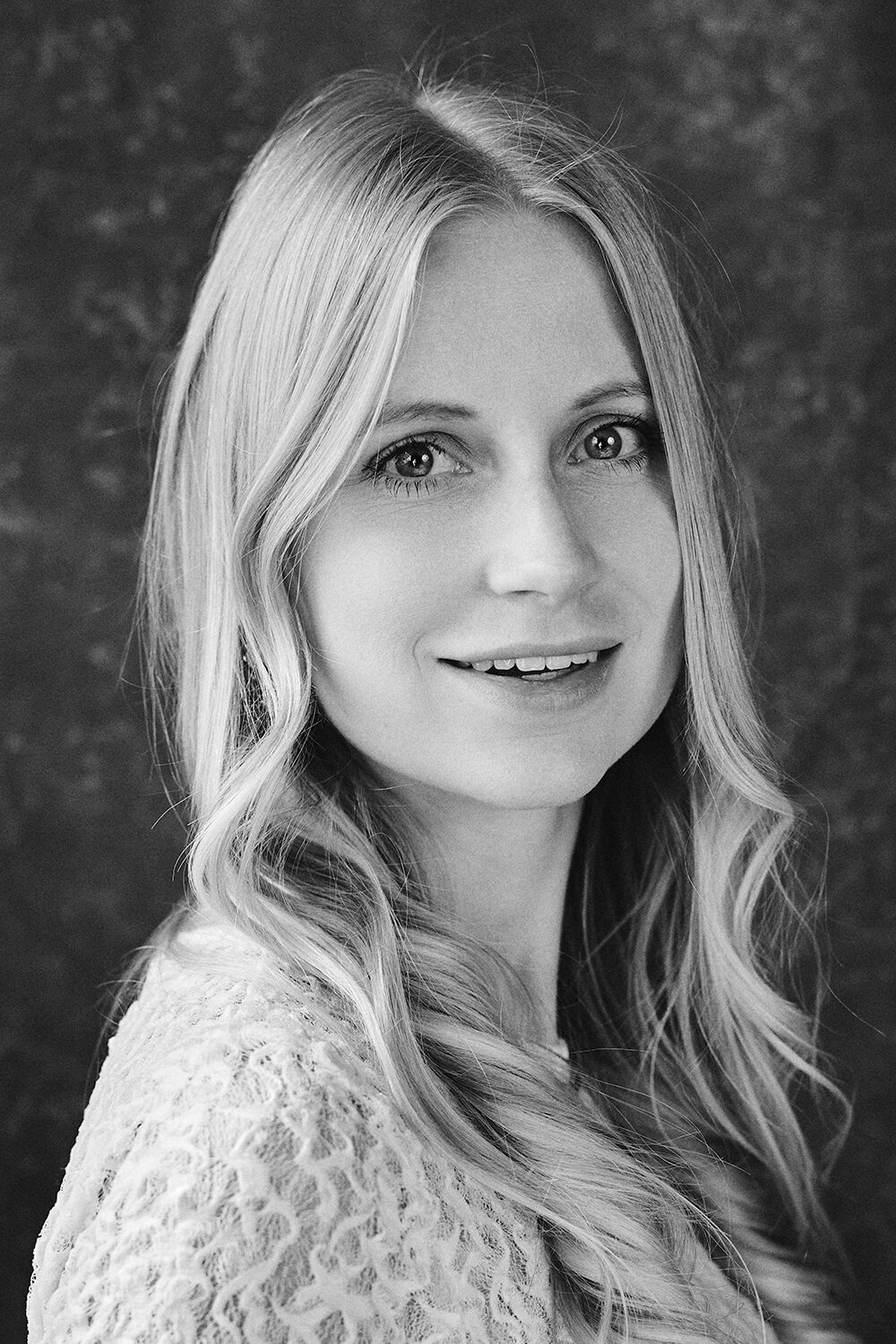 Schwarz-Weiß-Portrait einer lächelnden blonden Frau in Spitzentop vor dunklem, strukturiertem Hintergrund.