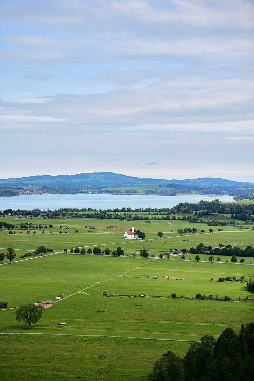 Blick von oben über weite grüne Wiesen auf eine kleine weiße Kirche und einen See im Allgäu, dahinter bewaldete Hügel.