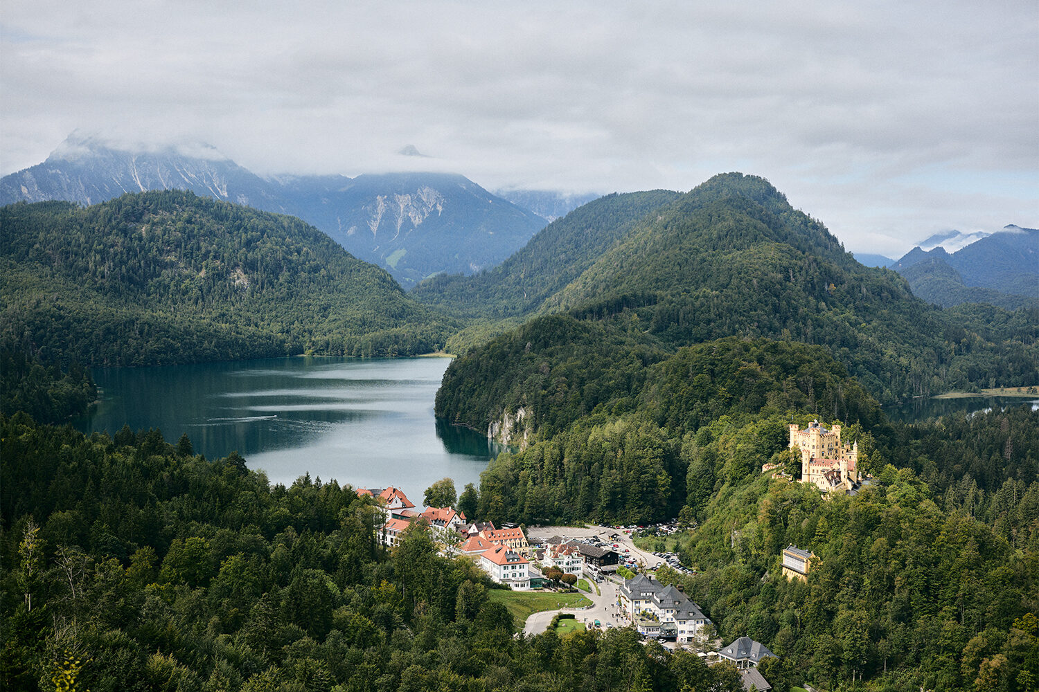 Blick auf den Alpsee mit Schloss Hohenschwangau über dem Dorf, dahinter die bewaldeten Hänge der Allgäuer Alpen.