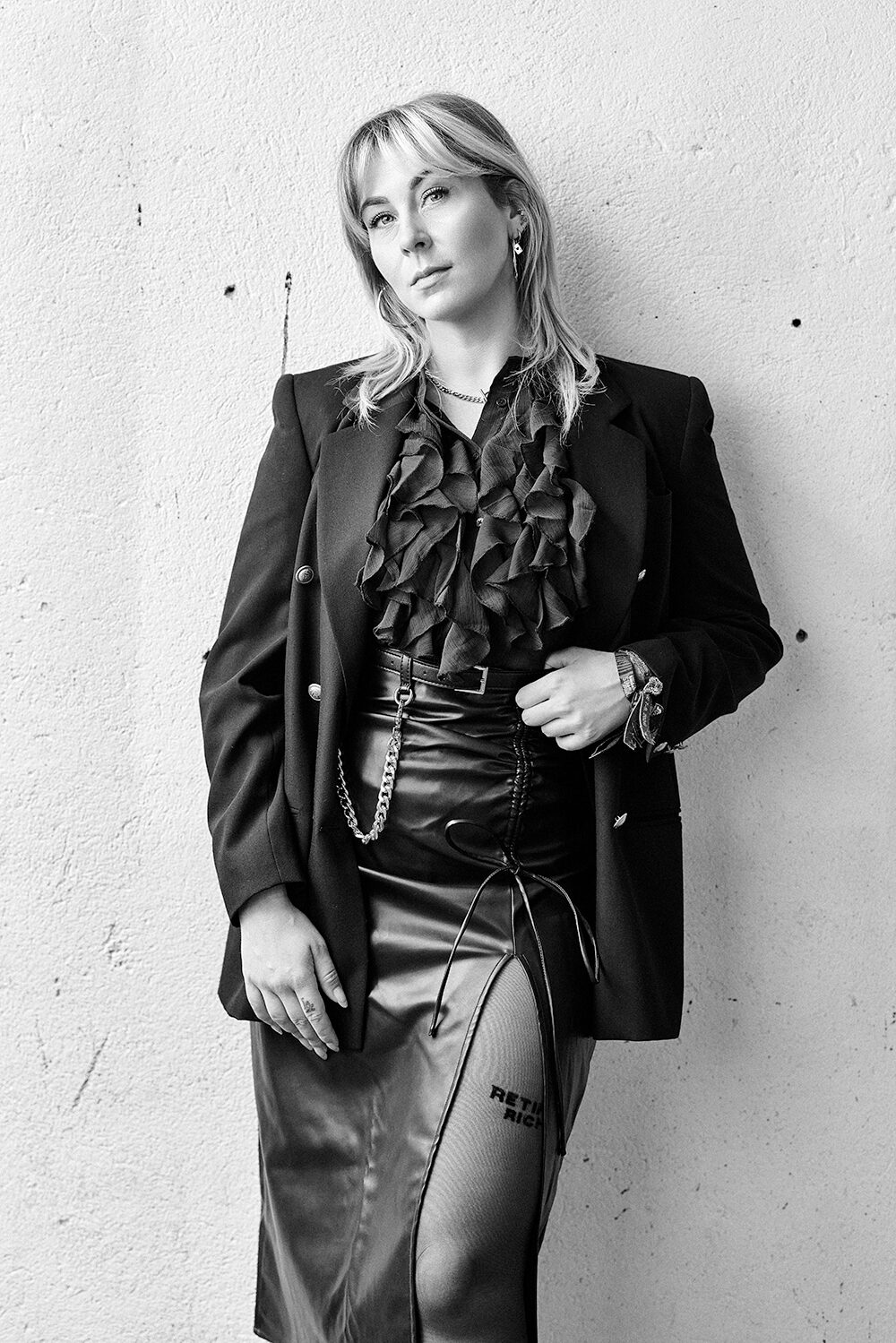 Schwarz-Weiß-Modeaufnahme: Frau im Oversize-Blazer über Rüschenbluse und Lederrock vor einer weißen Wand.