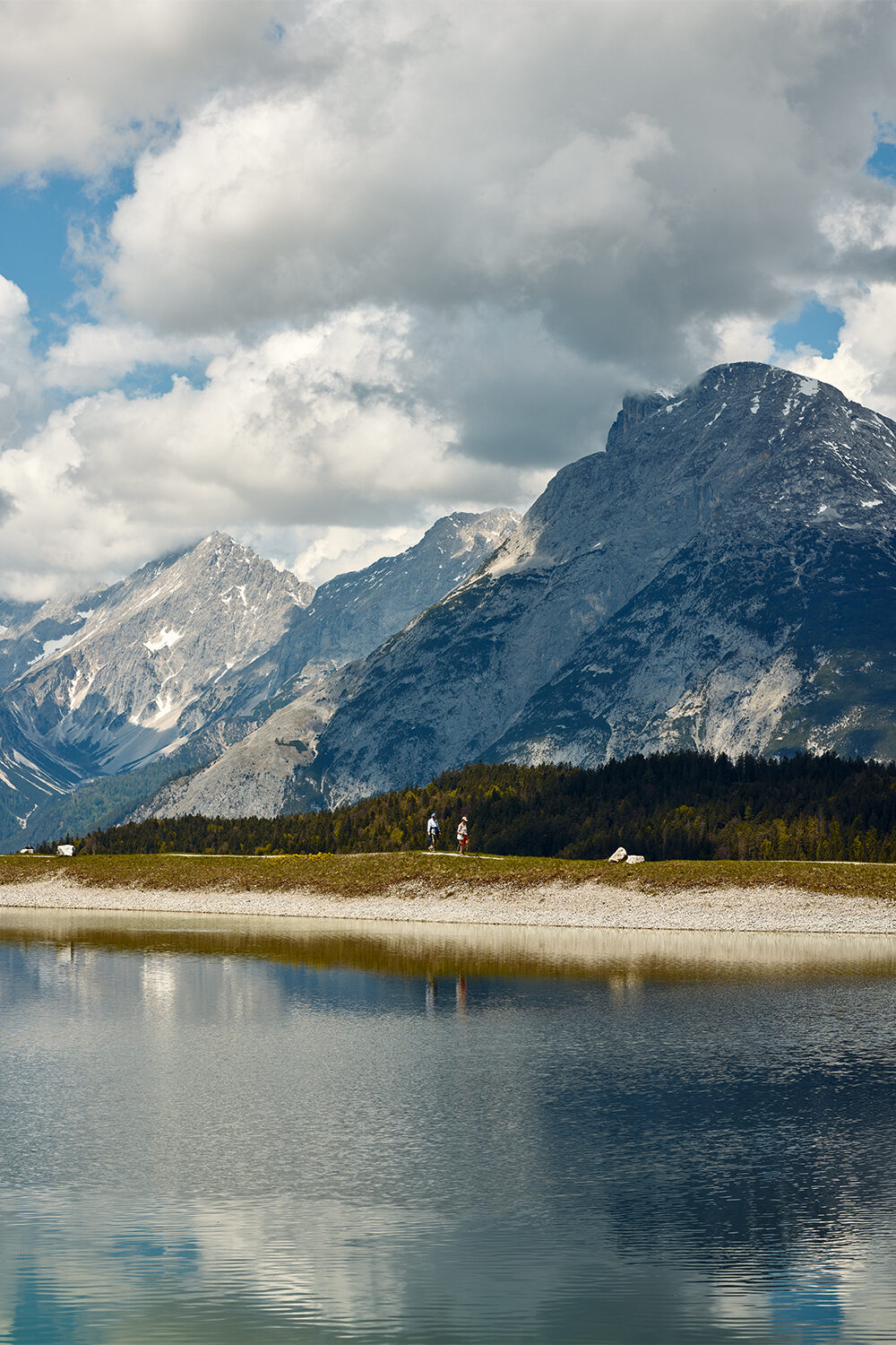 Schroffe Kalkgipfel spiegeln sich in einem Bergsee bei Seefeld in Tirol, am Ufer stehen zwei Wanderer.