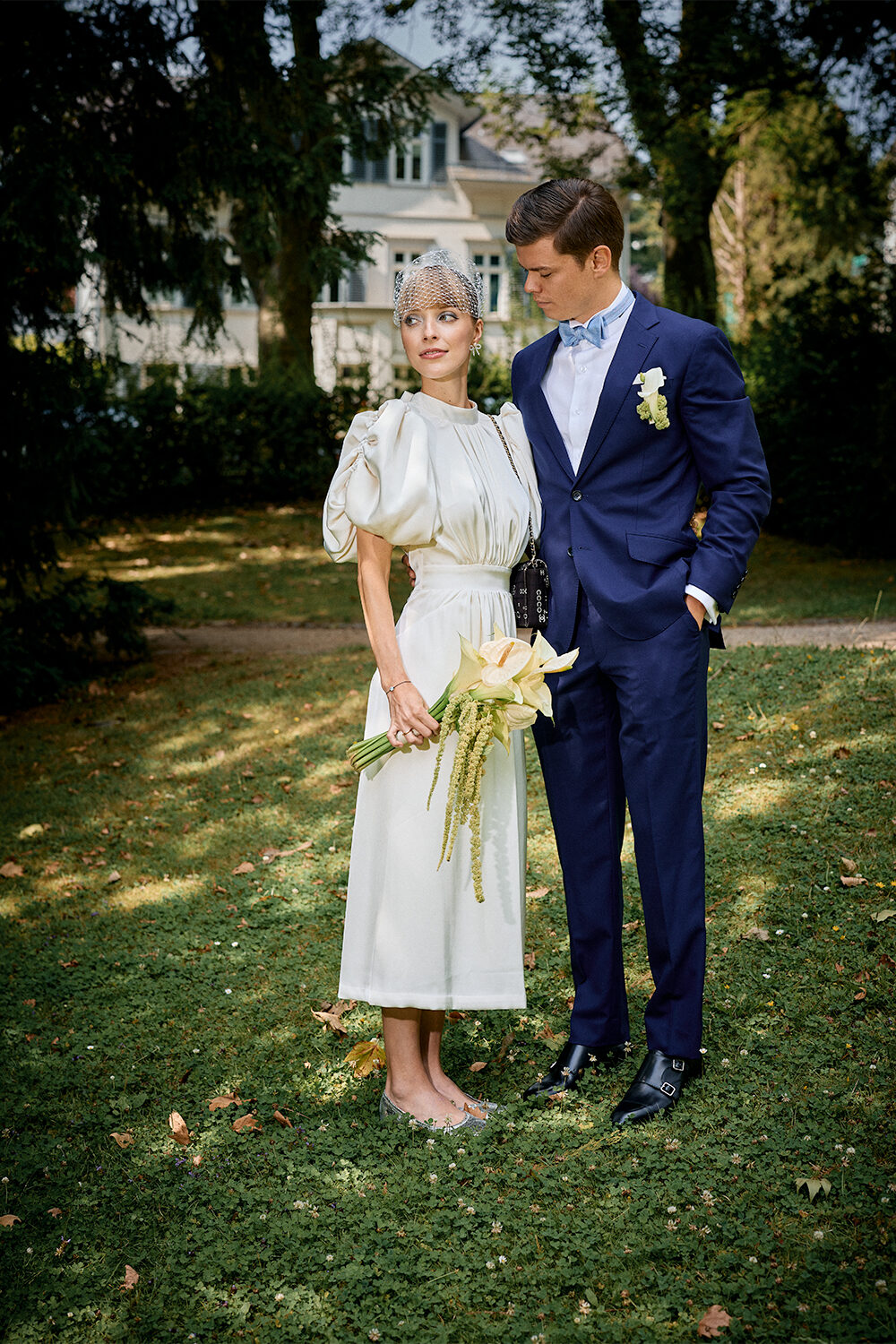 Brautpaar im Garten: die Braut im weißen Kleid mit Schleiertuch und Calla-Strauß, der Bräutigam im dunkelblauen Anzug mit Fliege.