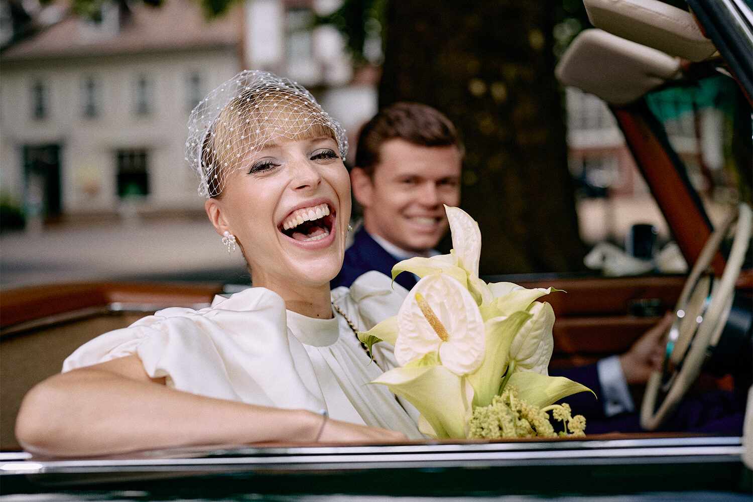 Lachende Braut mit Schleiertuch und Calla-Strauß im offenen Oldtimer, dahinter der Bräutigam.