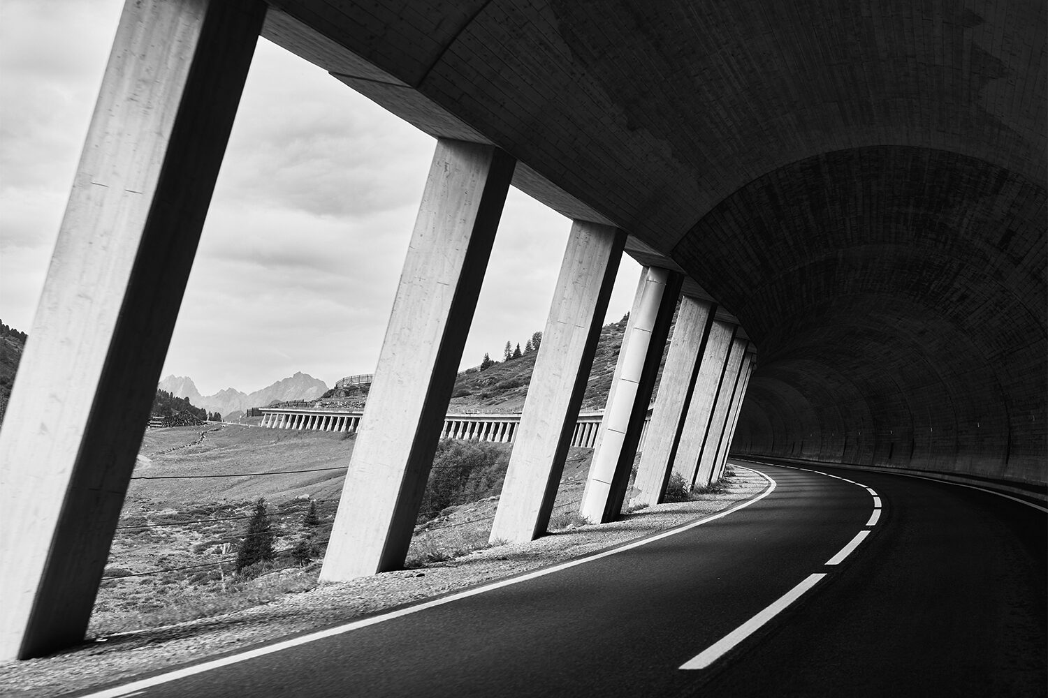 Schwarz-Weiß-Aufnahme: eine Straße führt durch eine Lawinengalerie, zwischen deren Betonpfeilern der Blick auf die Berge fällt.