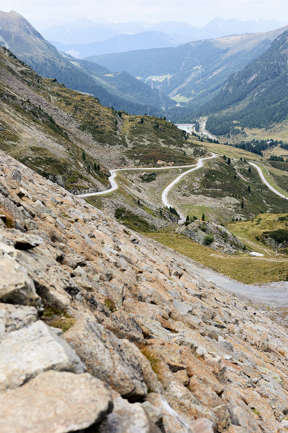 Blick über einen Geröllhang auf eine Passstraße, die sich in Serpentinen durch ein grünes Hochtal windet.