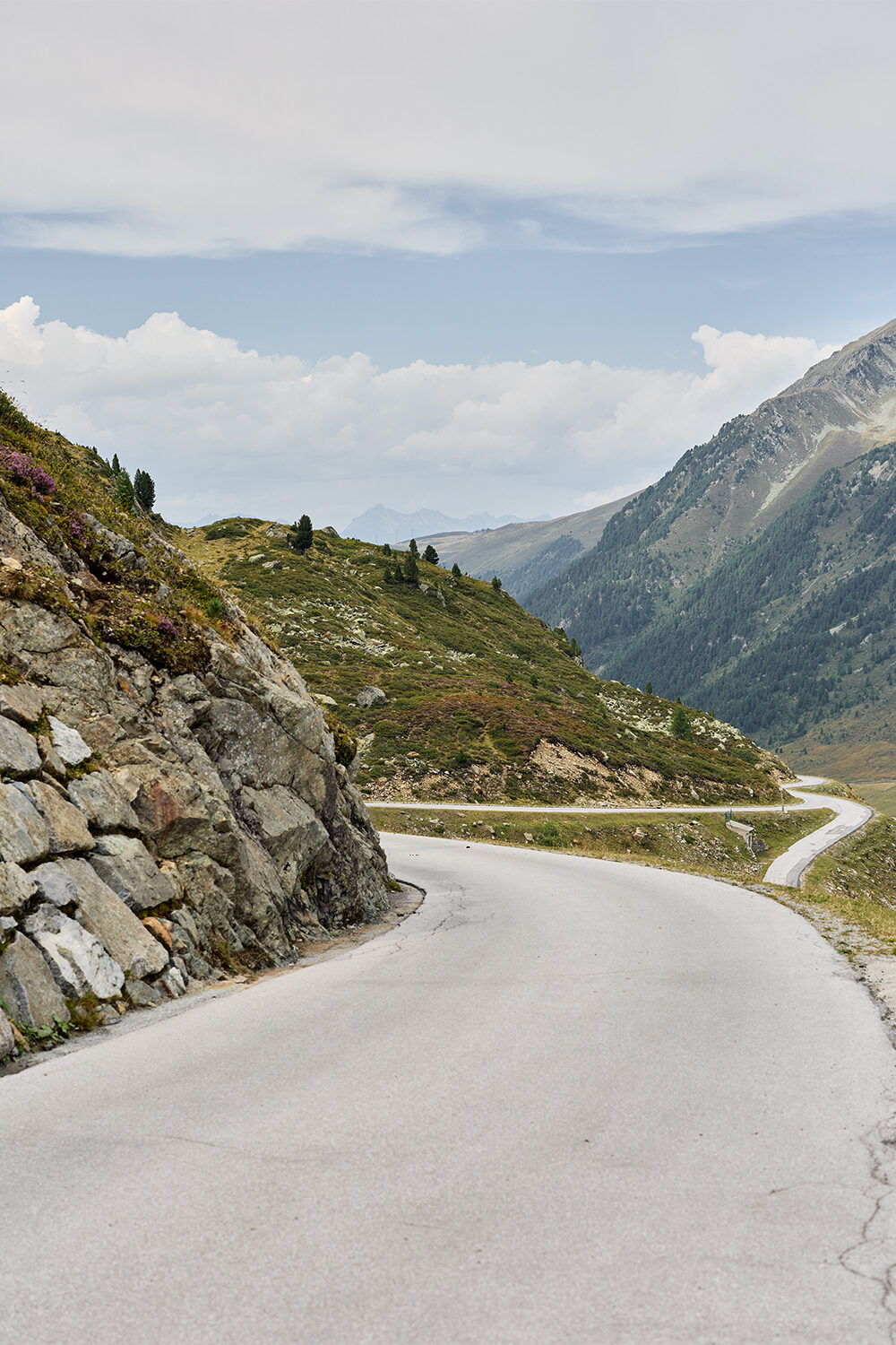 Eine Passstraße führt an einer Felswand vorbei in ein weites Hochtal, darüber ziehen Wolken.
