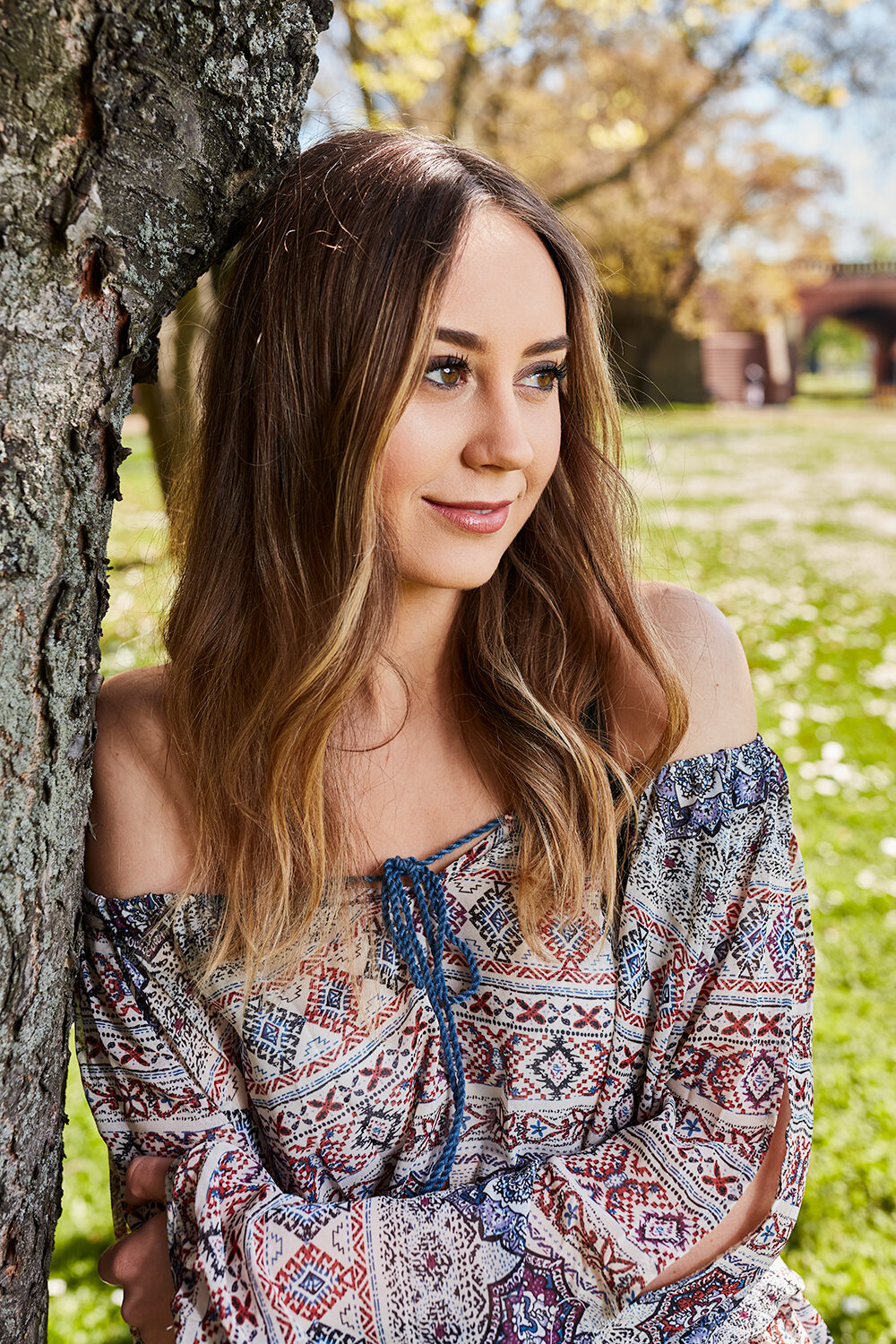 Lifestyle-Portrait einer brünetten Frau in Boho-Bluse, angelehnt an einen Baum im Park.