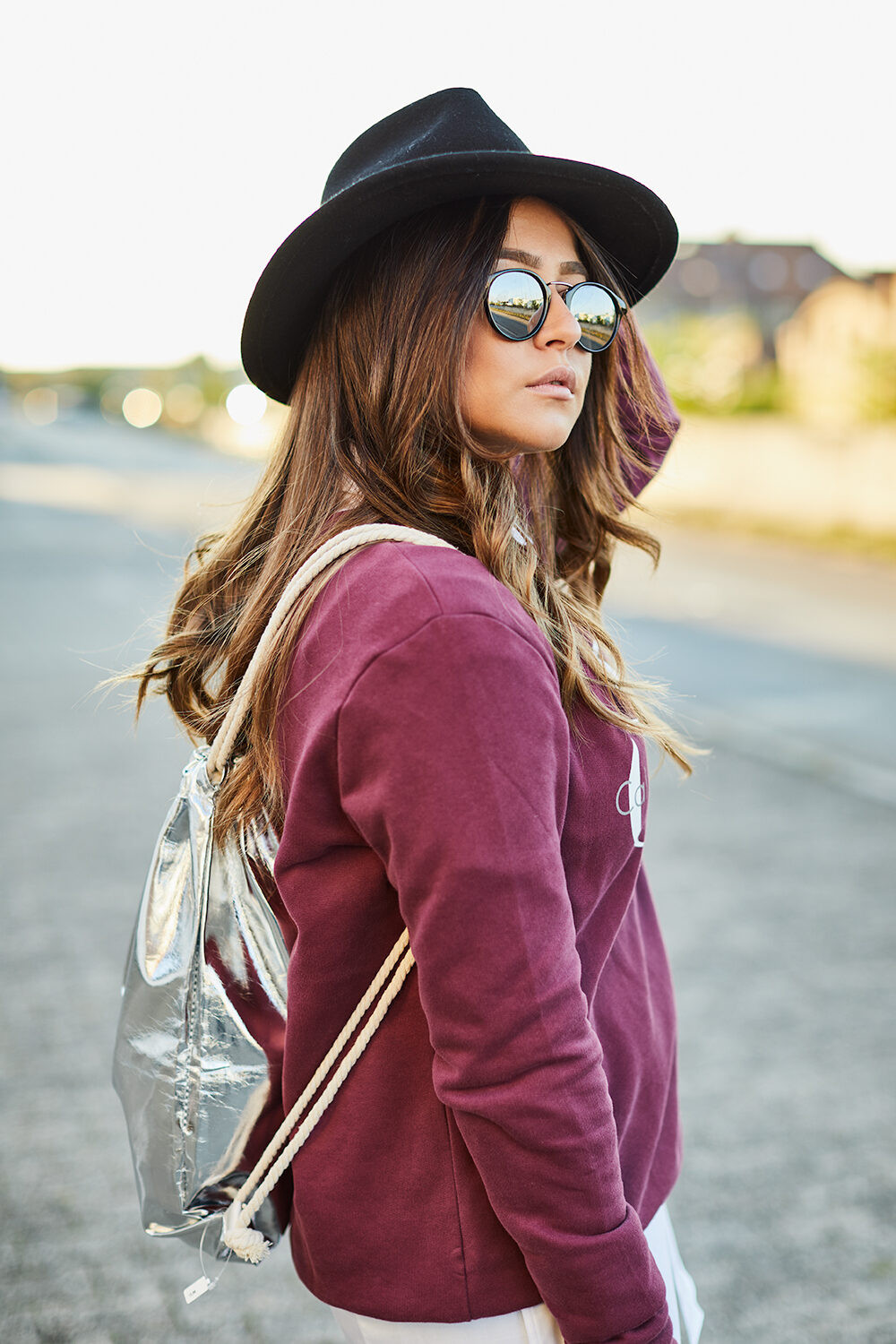 Brünette Frau mit schwarzem Hut, verspiegelter Sonnenbrille und Metallic-Rucksack auf einer Landstraße im Abendlicht.