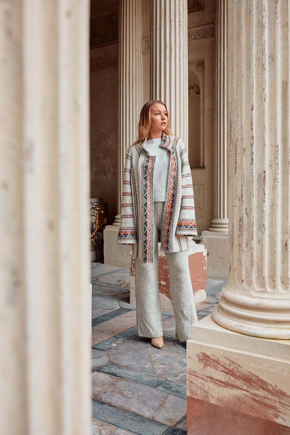 Blonde Frau in gemustertem Strickmantel zwischen Marmorsäulen einer prunkvollen Halle.