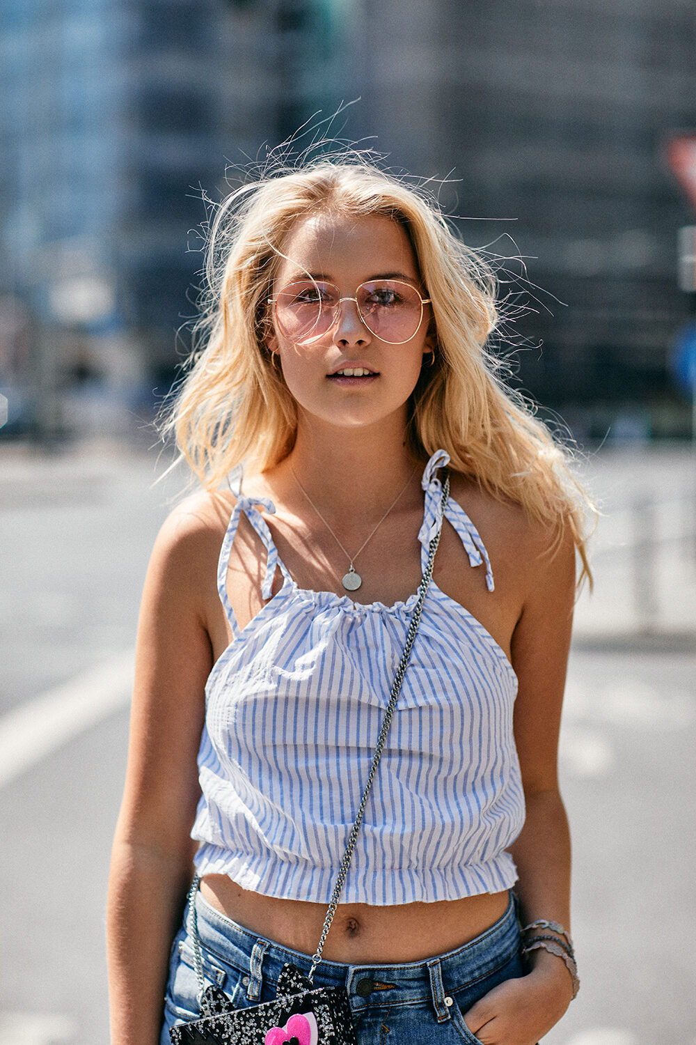 Streetstyle-Aufnahme einer jungen blonden Frau mit runder Sonnenbrille und gestreiftem Top auf einer sonnigen Stadtstraße.