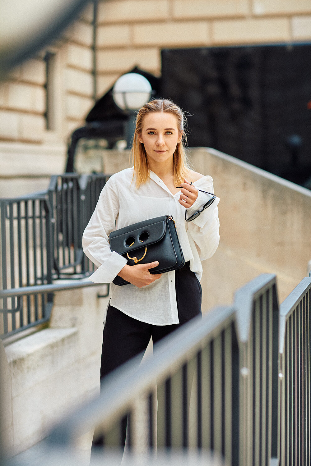 Blonde Frau in weißer Bluse und schwarzer Hose mit schwarzer Henkeltasche an einem Geländer in der Stadt.