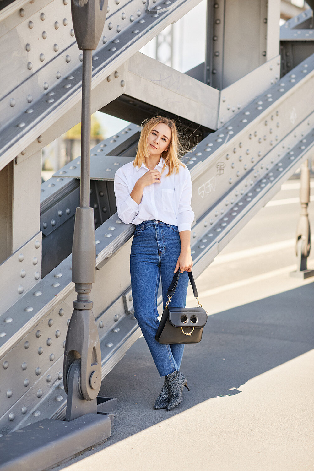 Blonde Frau in weißem Hemd und Jeans lehnt an der Stahlkonstruktion einer Brücke.