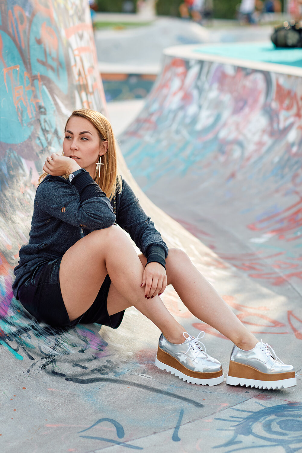 Blonde Frau in grauem Sweatshirt und silbernen Plateauschuhen sitzt in einer mit Graffiti bemalten Skatepark-Rampe.
