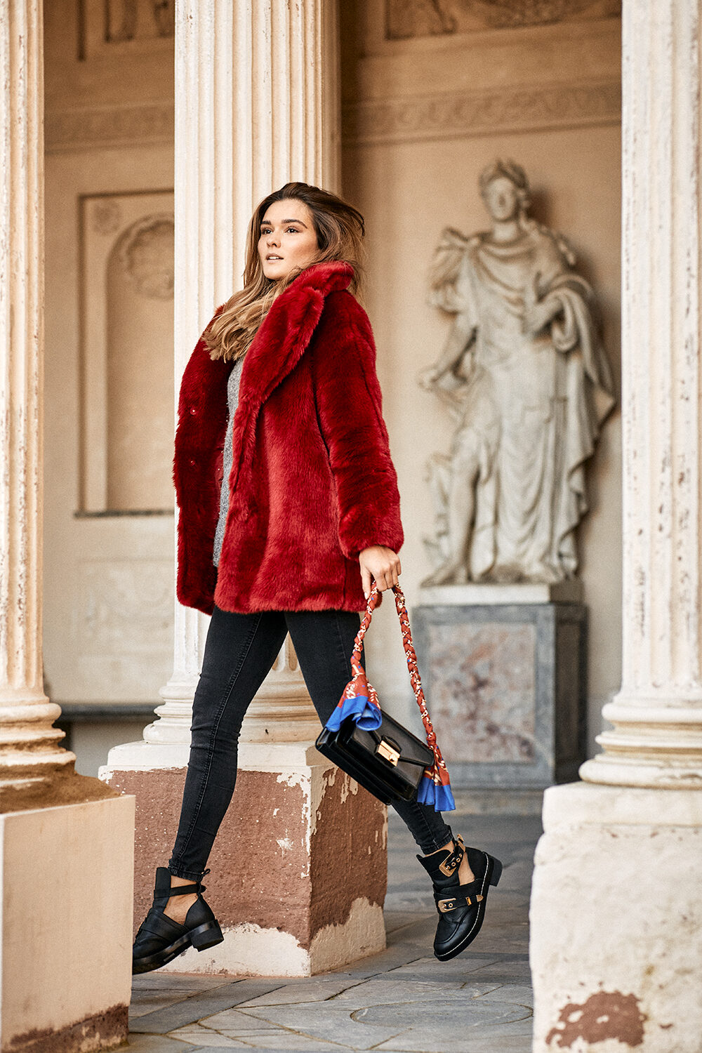 Die Fashion- und Lifestyle-Bloggerin Lea Endys im roten Felljacken-Mantel geht durch den Säulengang eines historischen Gebäudes mit Statue.