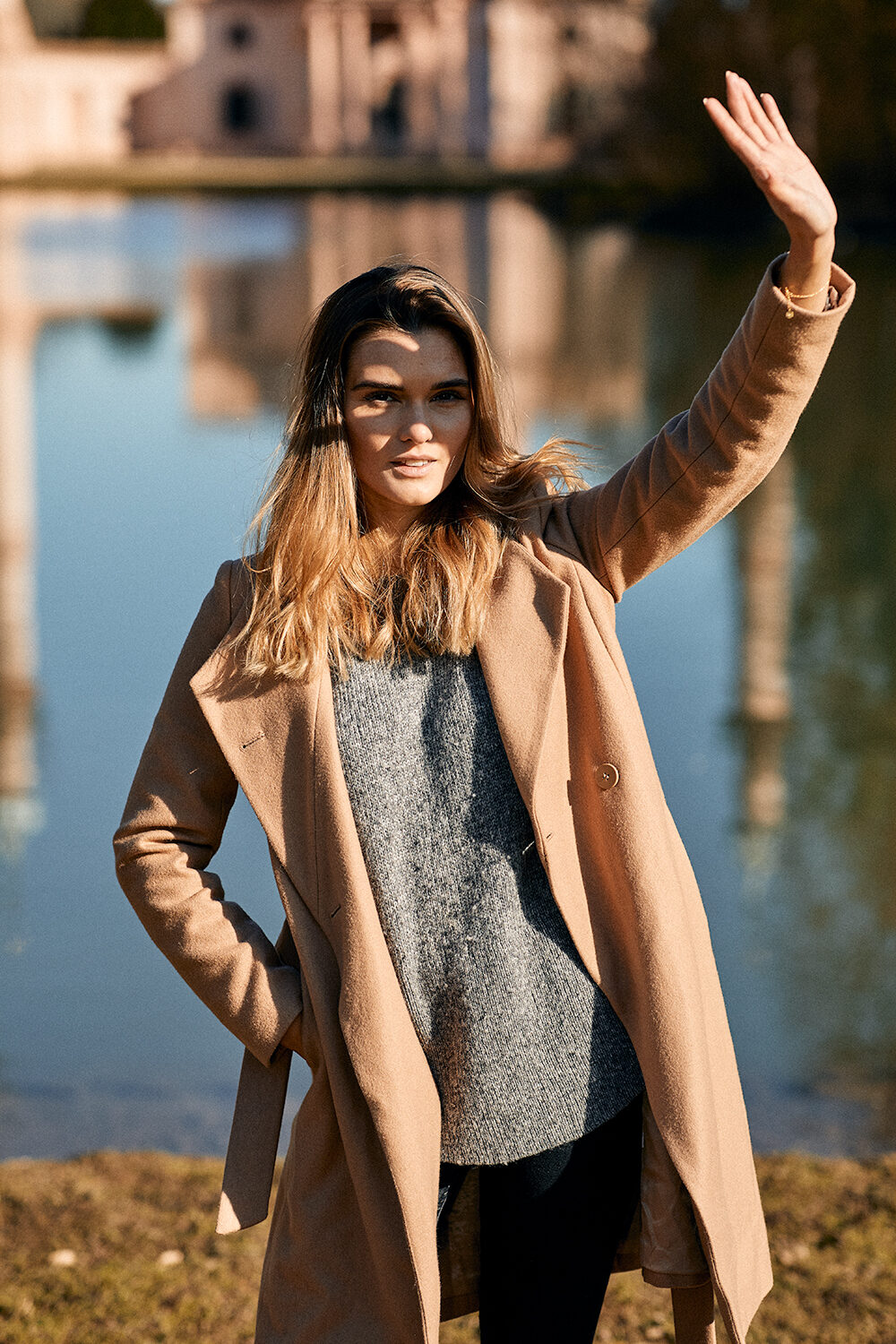 Lea Endys im Camel-Mantel über grauem Strickpullover winkt an einem Weiher, im Hintergrund ein historisches Gebäude.