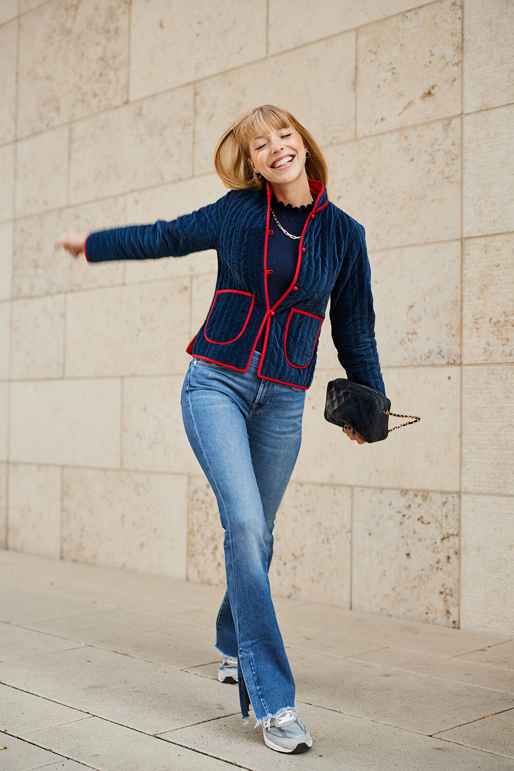Lachende Frau in marineblauer Strickjacke und Schlagjeans tanzt vor einer hellen Steinwand.