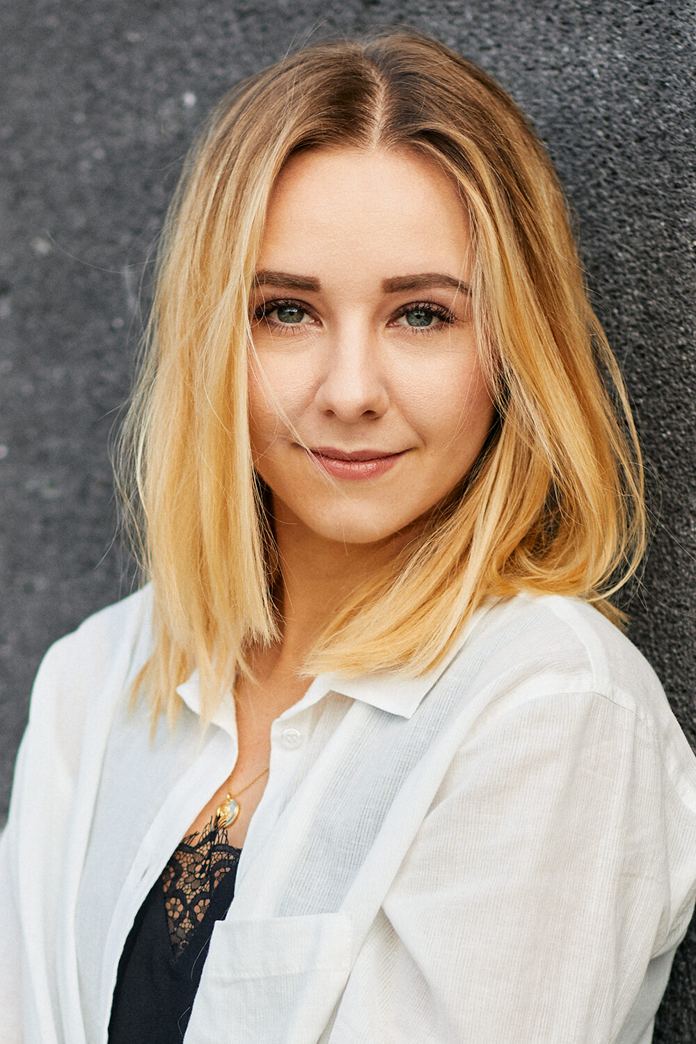 Portrait einer jungen blonden Frau im weißen Hemd vor einer grauen Wand.