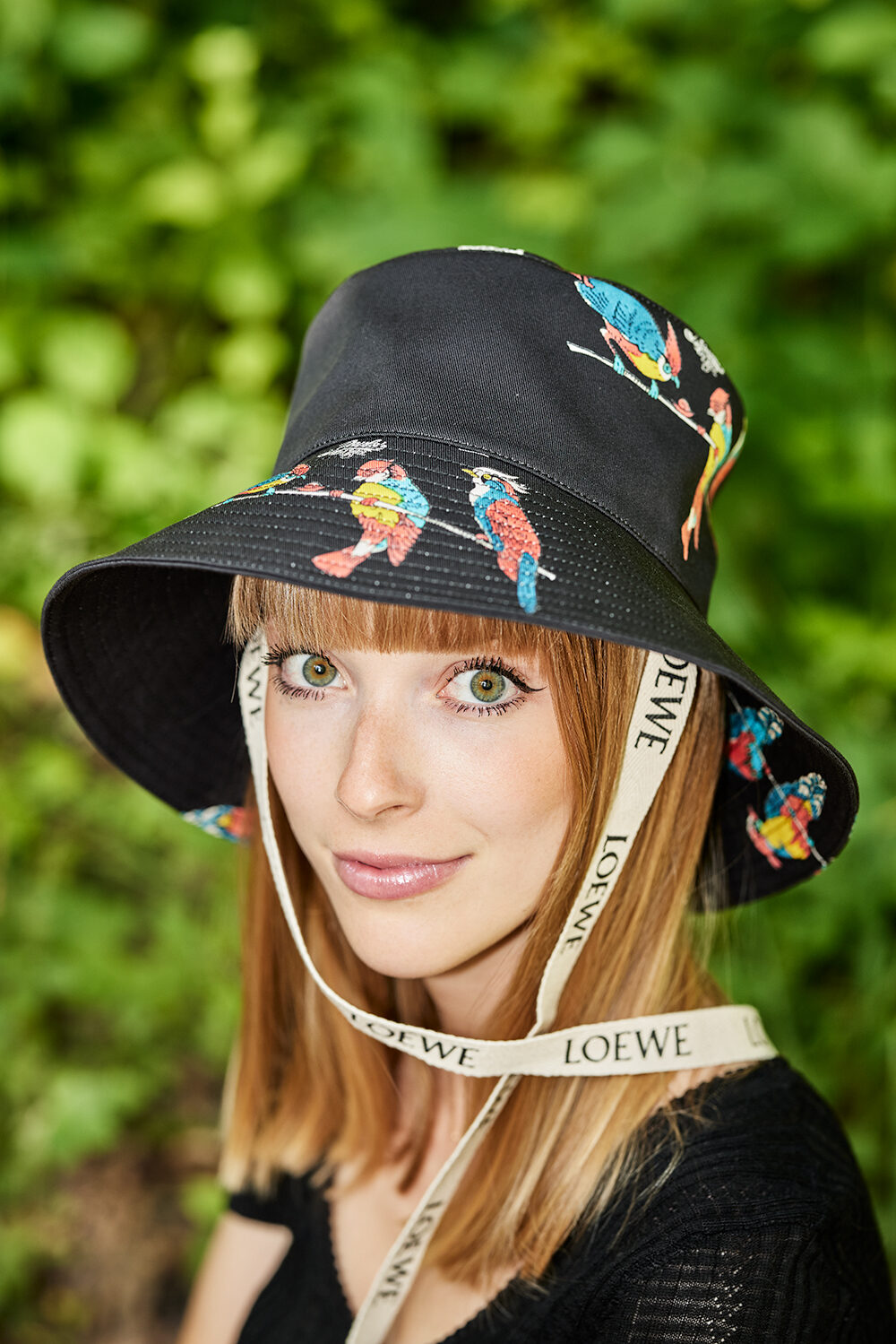 Portrait einer jungen Frau mit gemustertem Loewe-Bucket-Hat vor grünem Laub.