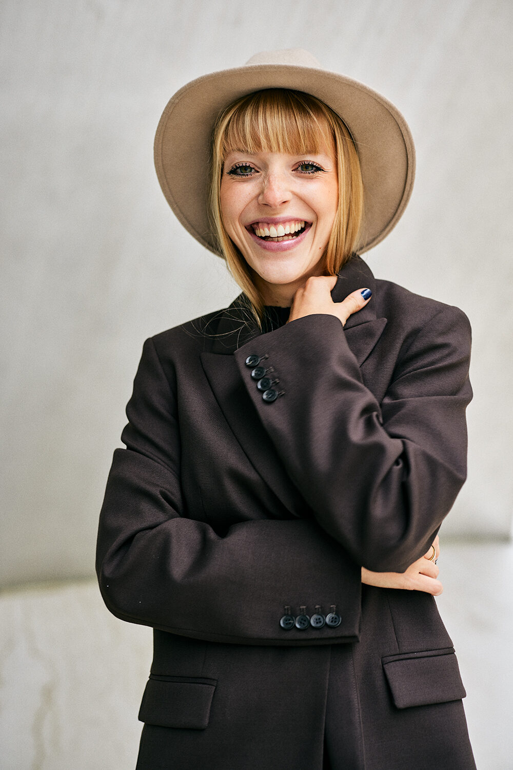 Portrait einer lachenden jungen Frau mit Filzhut und Blazer vor heller Wand.