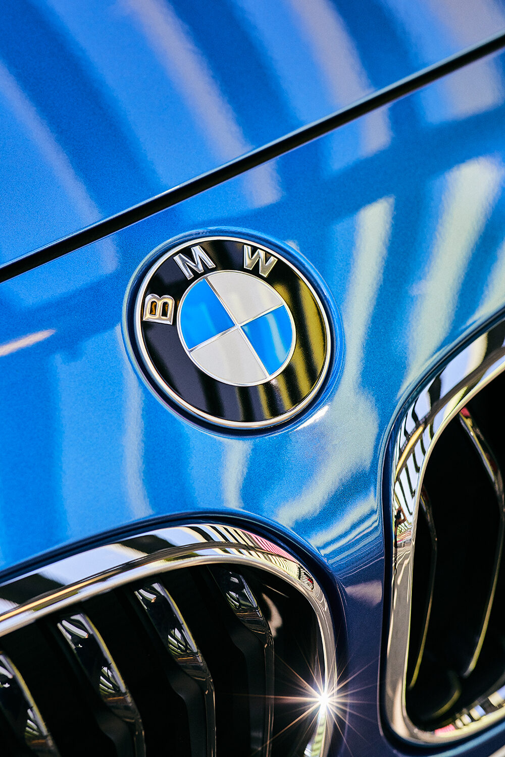 Detailaufnahme des BMW-Logos auf der Motorhaube eines estorilblauen BMW M140i xDrive mit den Nieren im Anschnitt.