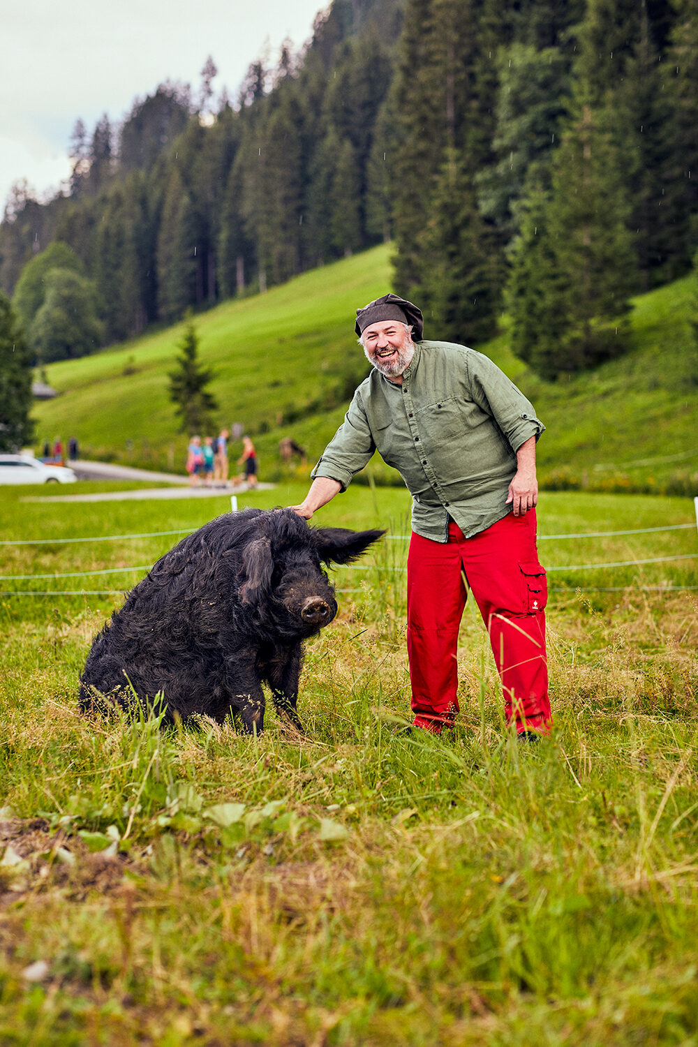 Vorarlberger Haubenkoch Jeremias Riezler lacht neben einem seiner schwarzen Weideschweine auf einer Bergwiese im Kleinwalsertal.