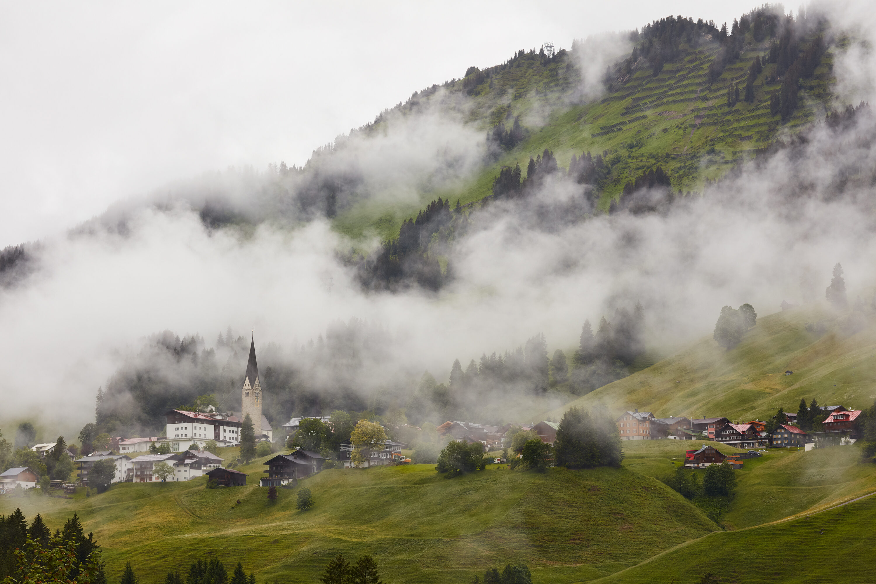 Tiefhängende Nebelschwaden verhüllen den Berghang über Kirchturm und Häusern von Mittelberg im Kleinwalsertal.