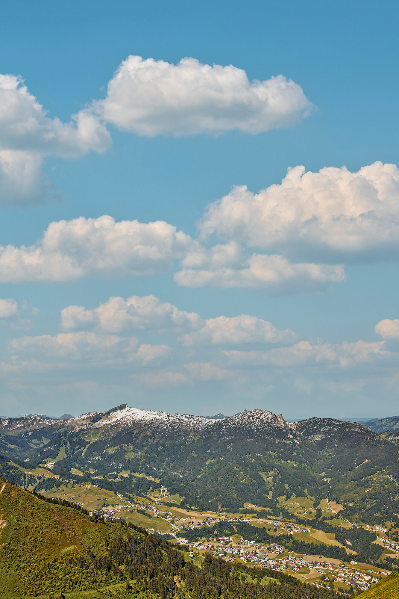 Blick über das Kleinwalsertal mit einem Dorf im Tal und schneebedeckten Gipfeln unter Schönwetterwolken.