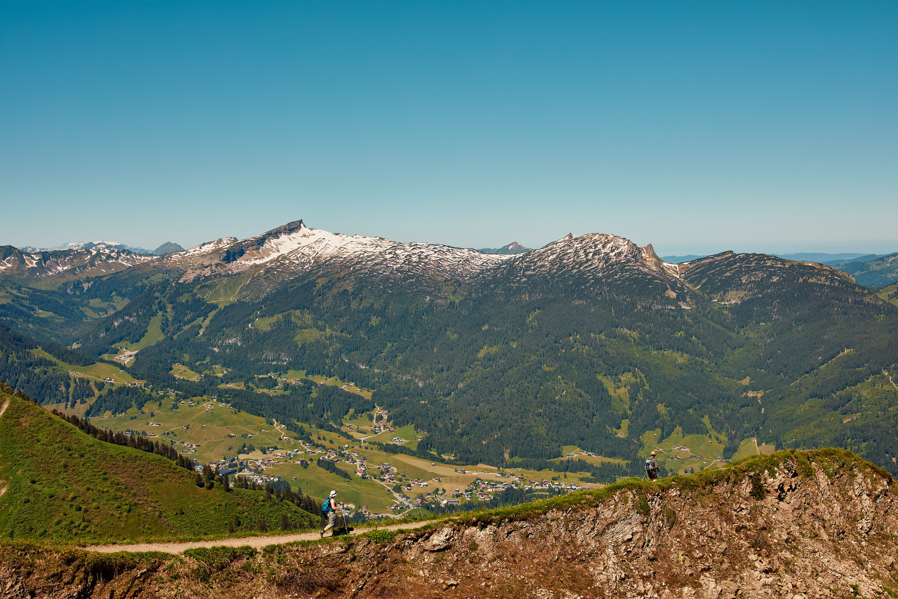 Panoramablick über das Kleinwalsertal mit Dorf im Tal, schneebedeckten Bergen und Wanderern auf dem Höhenweg.