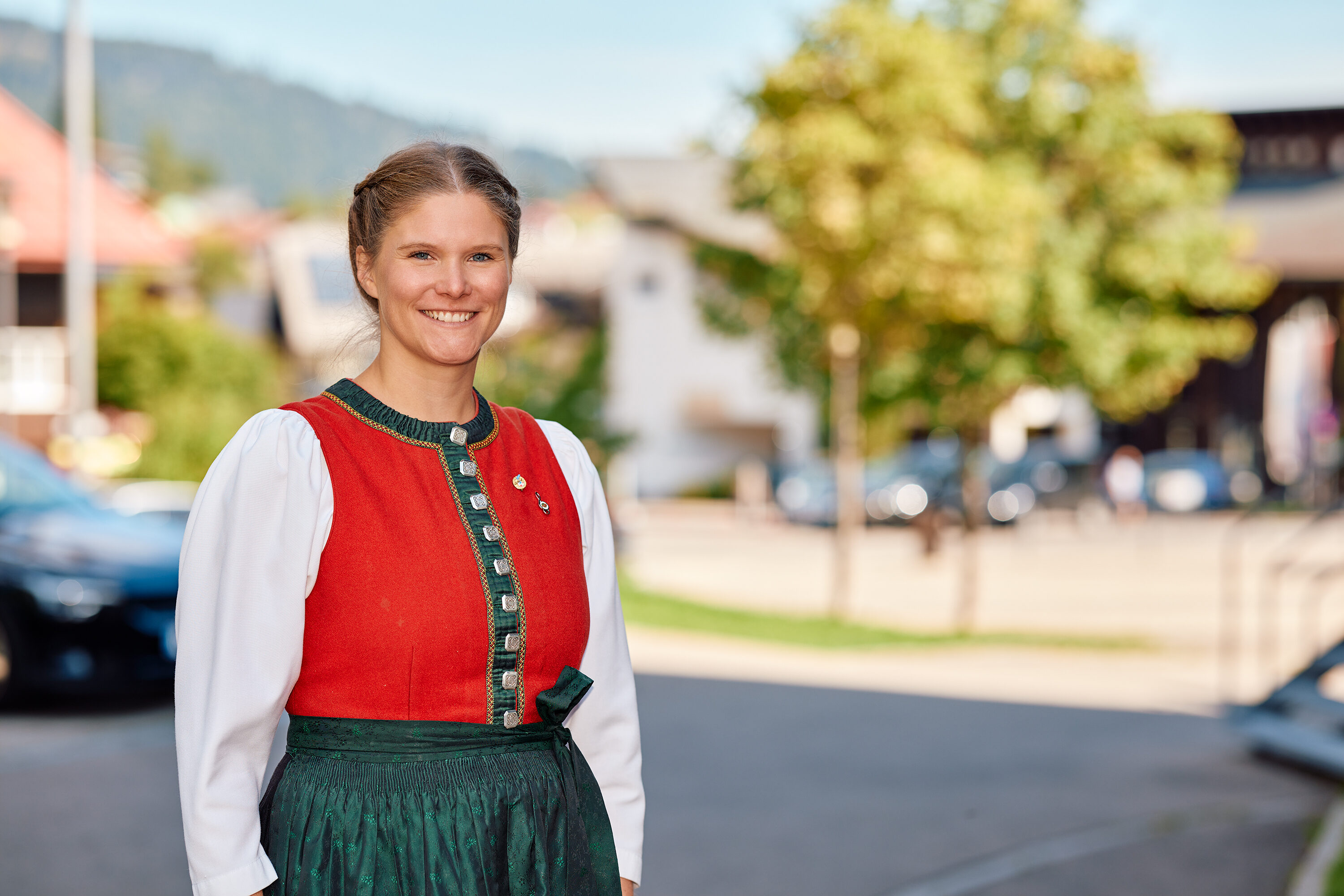 Katrin Berchtold in traditioneller Walser Tracht mit rotem Mieder und grüner Schürze steht lächelnd an einer Dorfstraße im Kleinwalsertal.